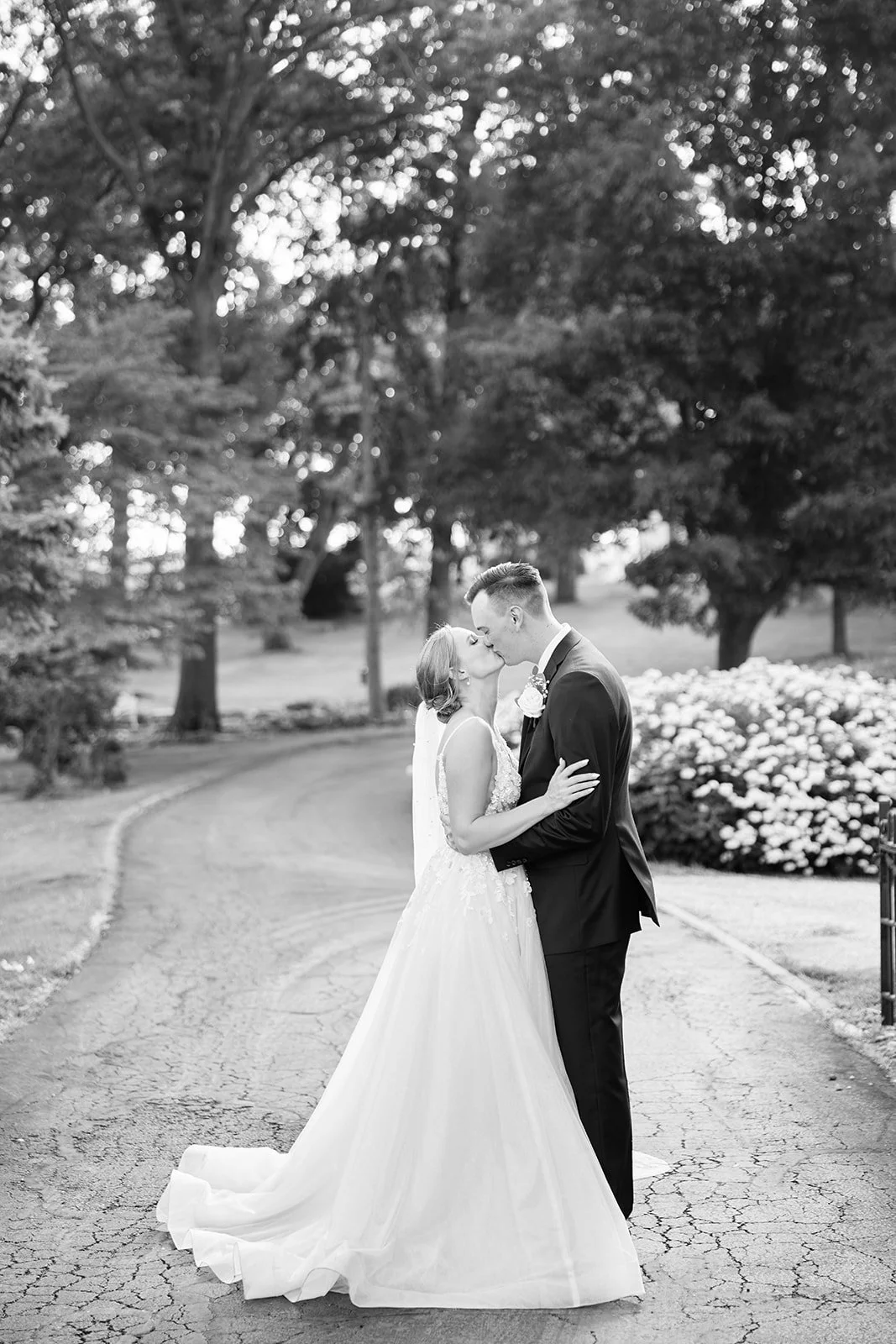 A bride and groom share a kiss outdoors on a wedding day, surrounded by trees and flowers.