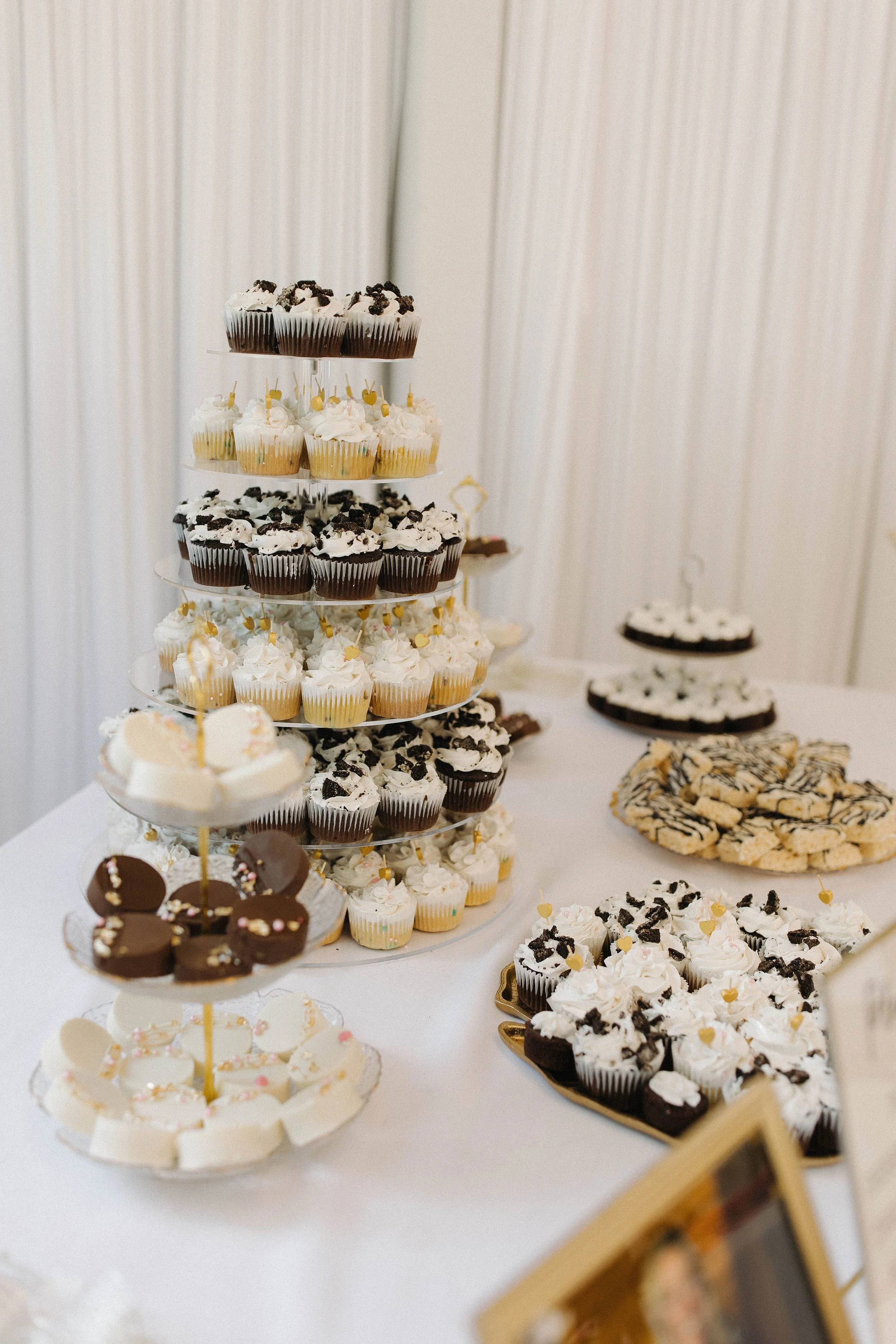 A dessert table with a tiered cupcake stand filled with cream and chocolate cupcakes decorated with Oreo cookies, alongside various cookies and other desserts on plates.