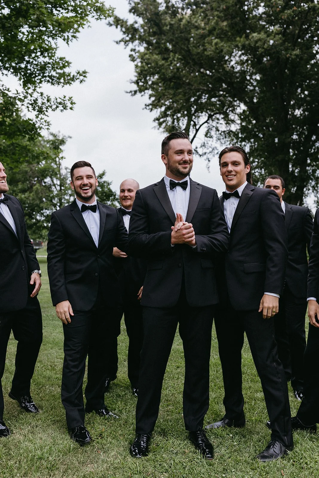 Group of men dressed in black tuxedos with bow ties, standing on grass outdoors, smiling and posing for a photo.