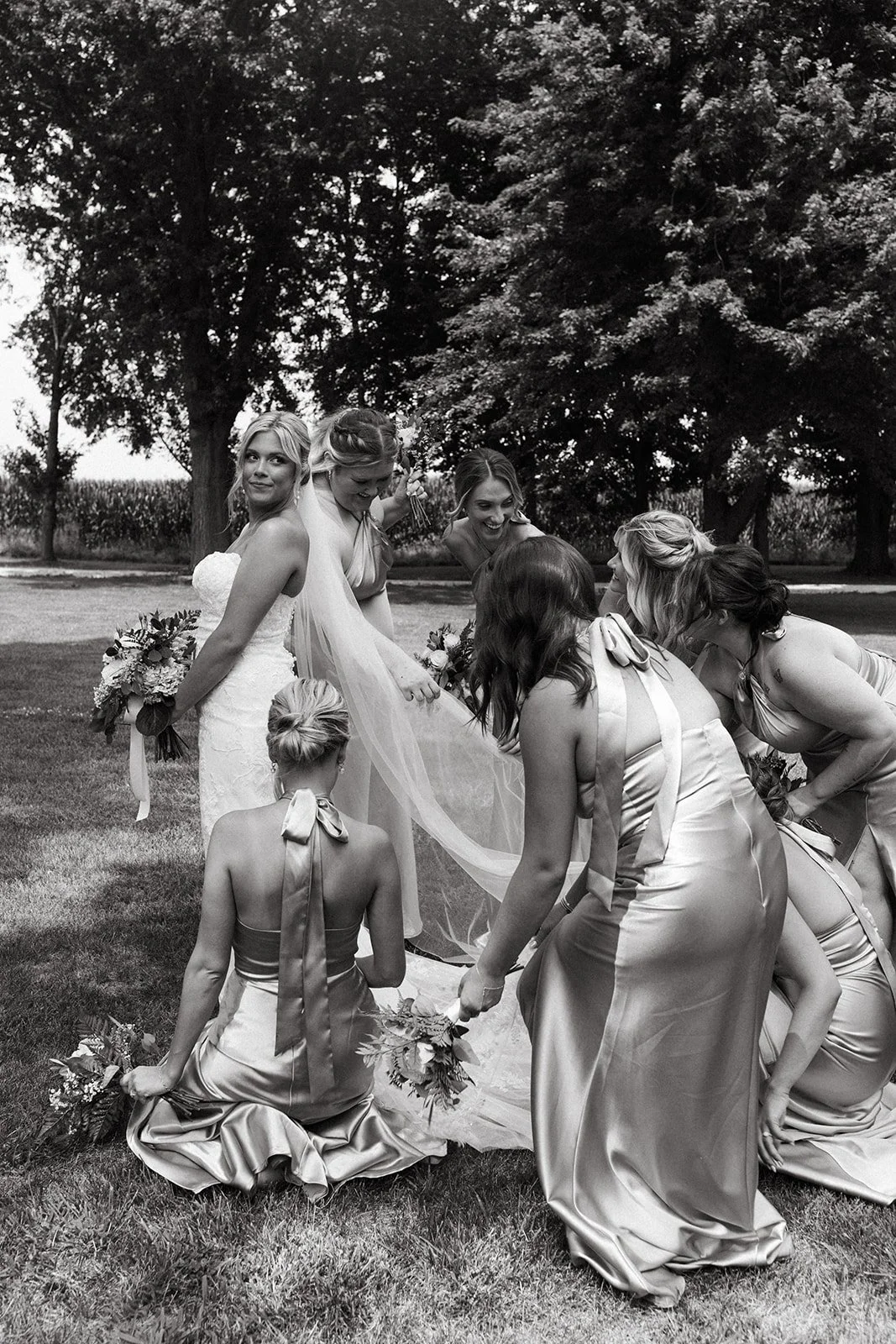 A group of women, including a bride in a white wedding dress, and her bridesmaids in satin dresses, outdoors on grass with trees in the background, preparing or celebrating a wedding.
