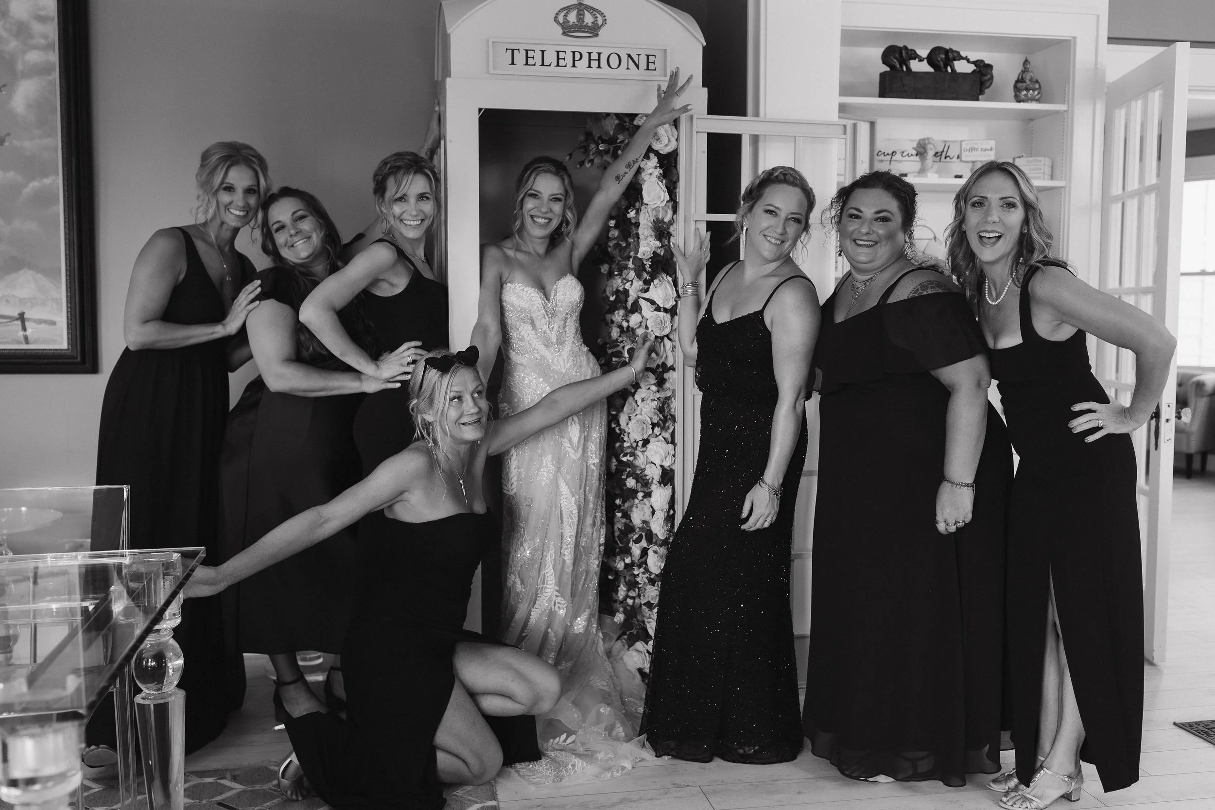 Group of women dressed in formal attire, posing in front of a floral arch and telephone booth, at a wedding celebration.