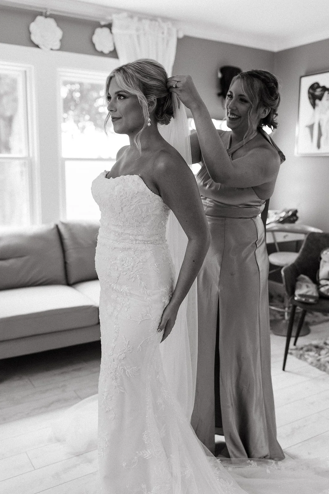 A bride in a strapless wedding dress with embroidery stands as another woman helps her put on a veil, in a cozy room with natural light from the windows.