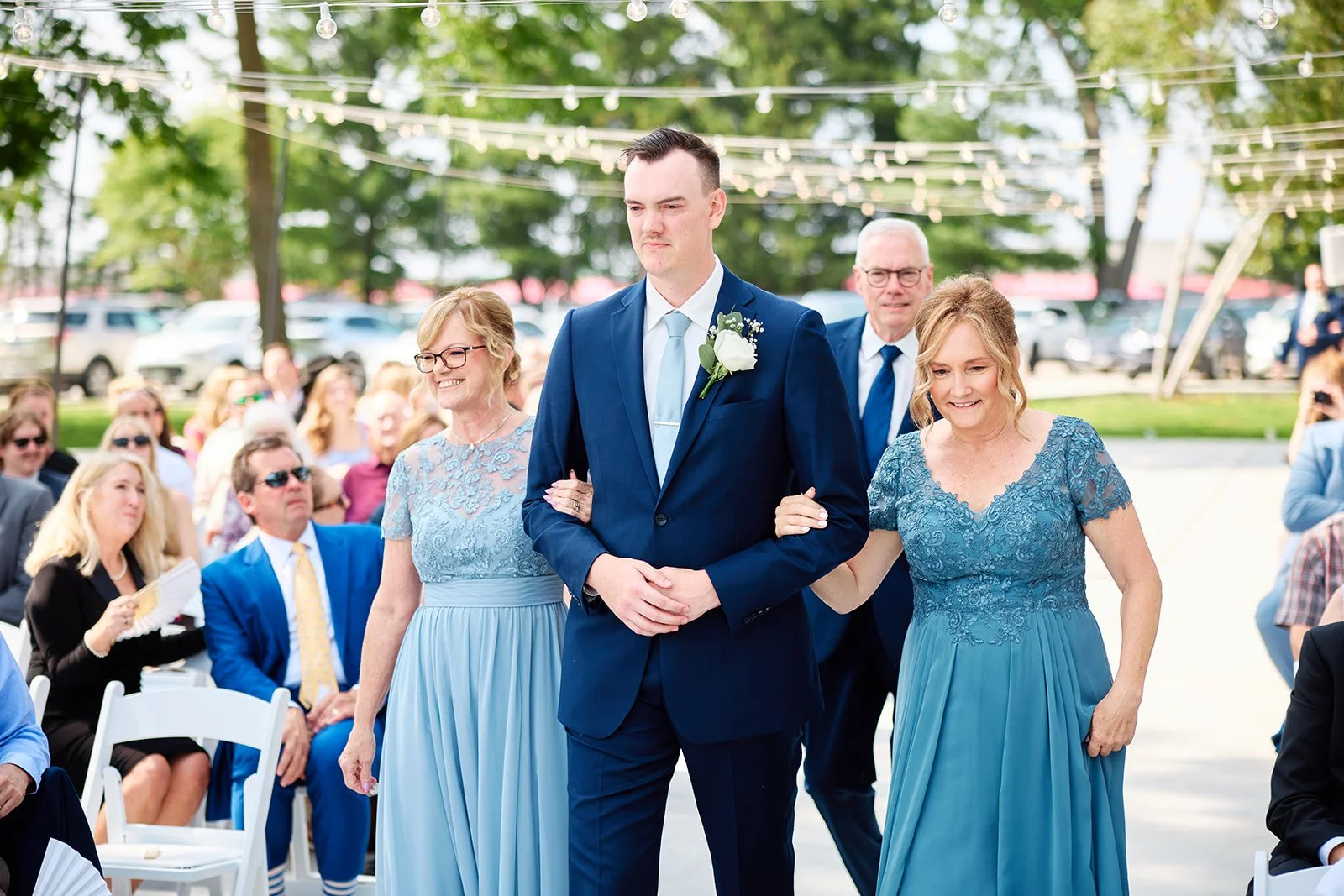 A groom in a navy blue suit walking down the aisle with his mother and father at an outdoor wedding. The groom has a white boutonniere, and the mother and father are dressed in matching blue dresses and suit with glasses. Guests are seated in the bac