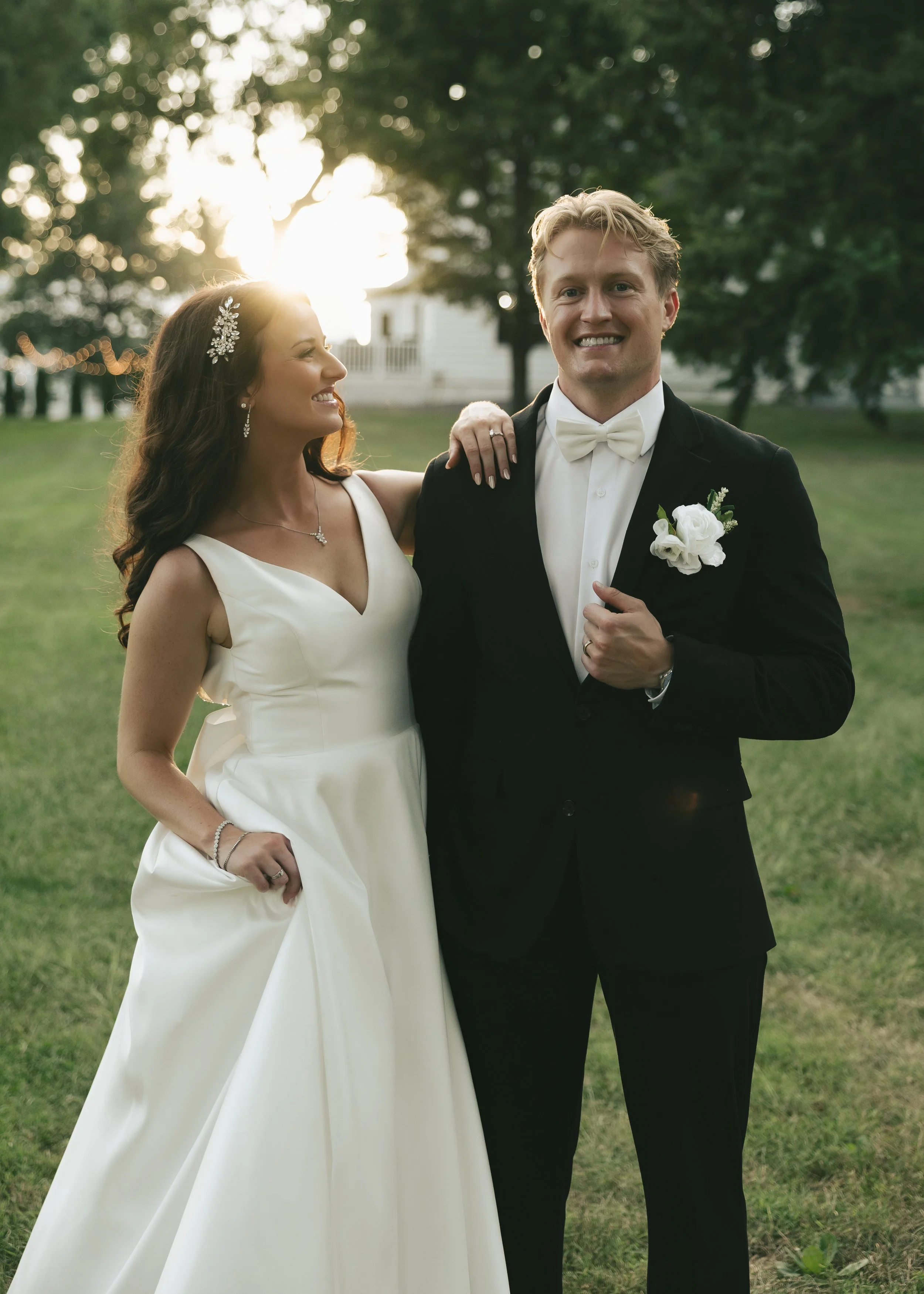 A bride and groom smiling outdoors during sunset, with the bride wearing a white dress and jewelry, and the groom in a black tuxedo with a white bow tie and boutonniere.