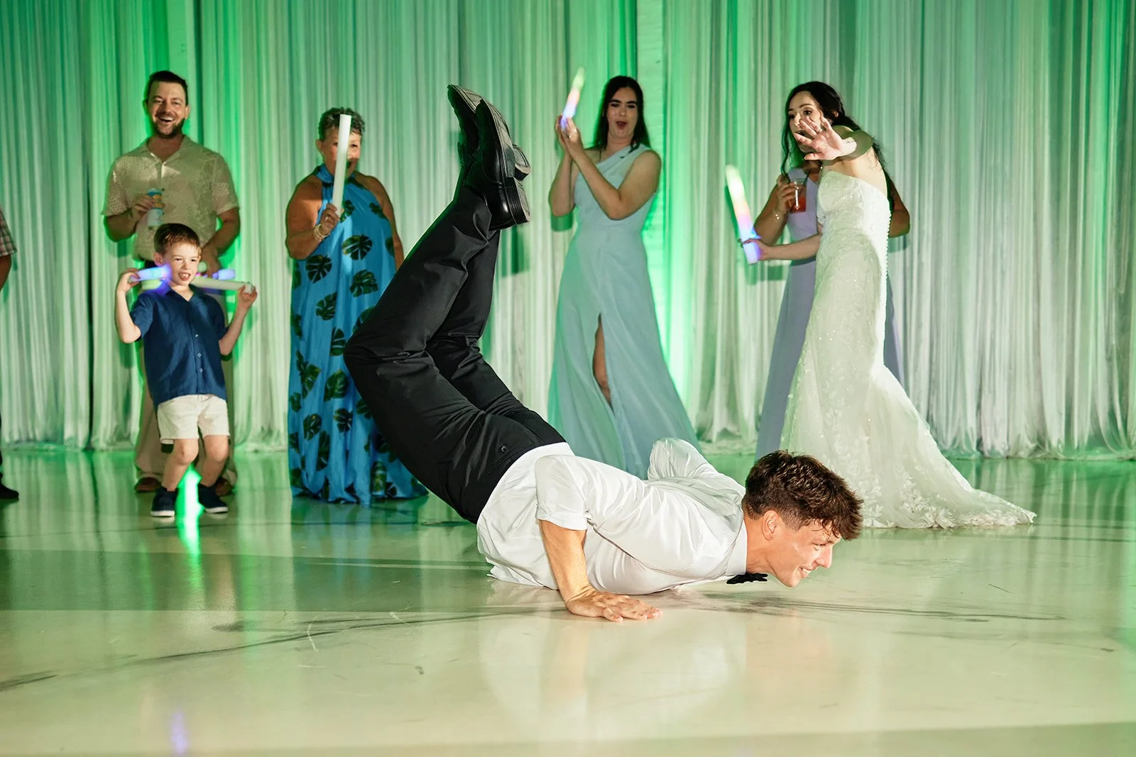 A groomsman dressed as a waiter performing a break dance move on the floor, with a group of people, including a bride and guests, watching and smiling in the background at a celebration or wedding reception.