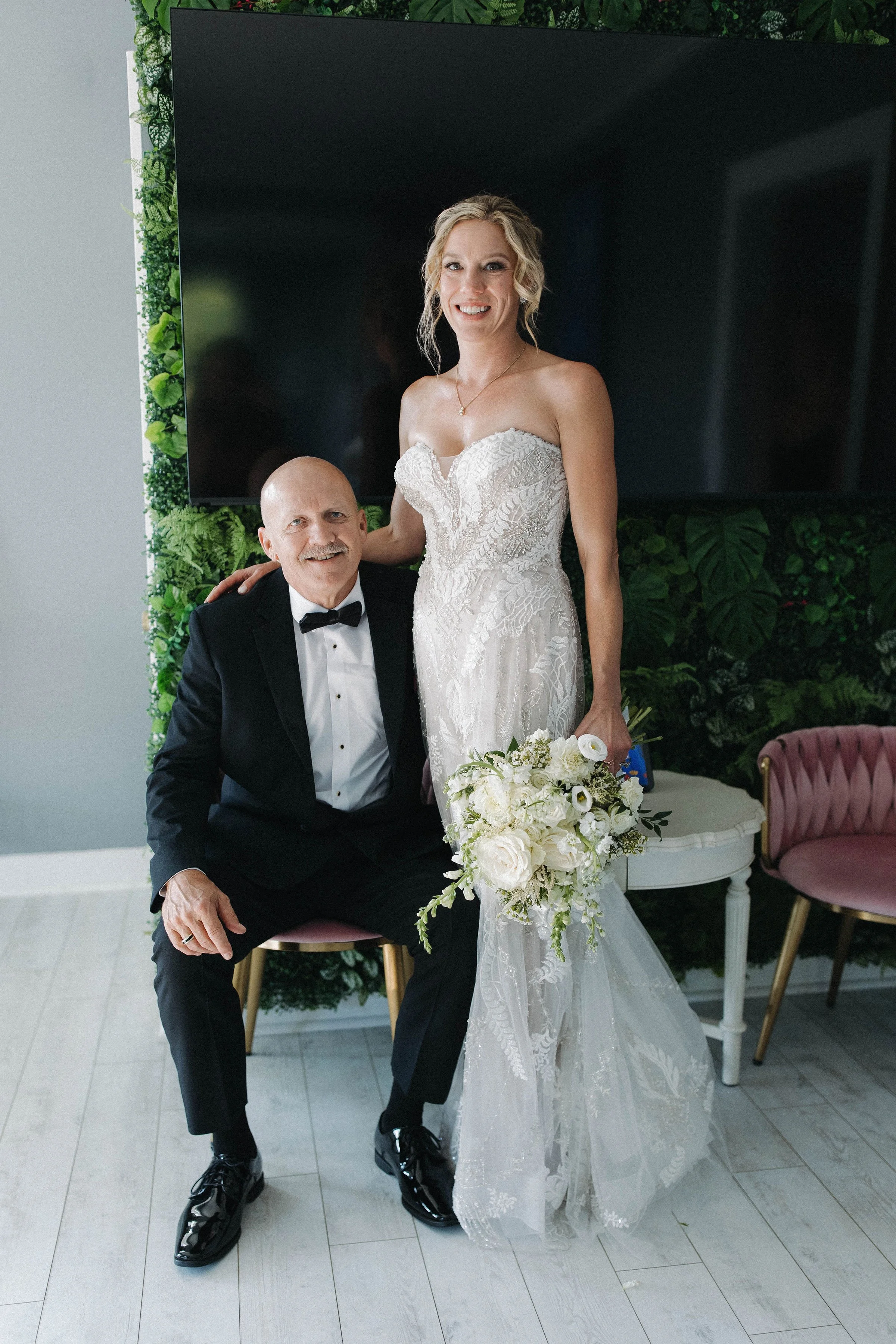 A bride in a strapless white wedding gown holding a bouquet of white flowers, standing beside a seated man in a tuxedo, both smiling at the camera indoors with greenery and a large black screen behind them.