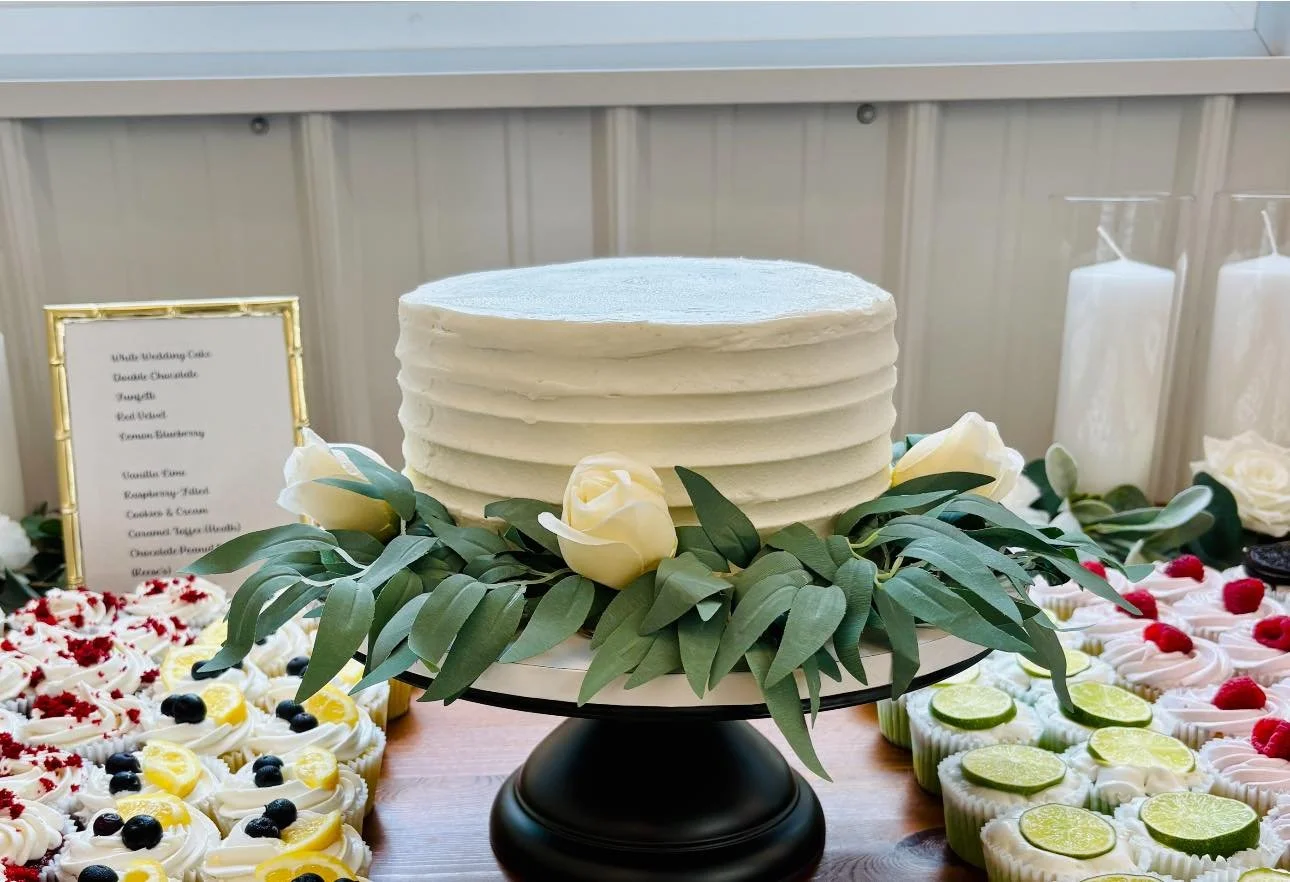 A white layered wedding cake decorated with white roses and green leaves, displayed on a black cake stand, surrounded by various cupcakes with fruit toppings and a menu sign in the background.