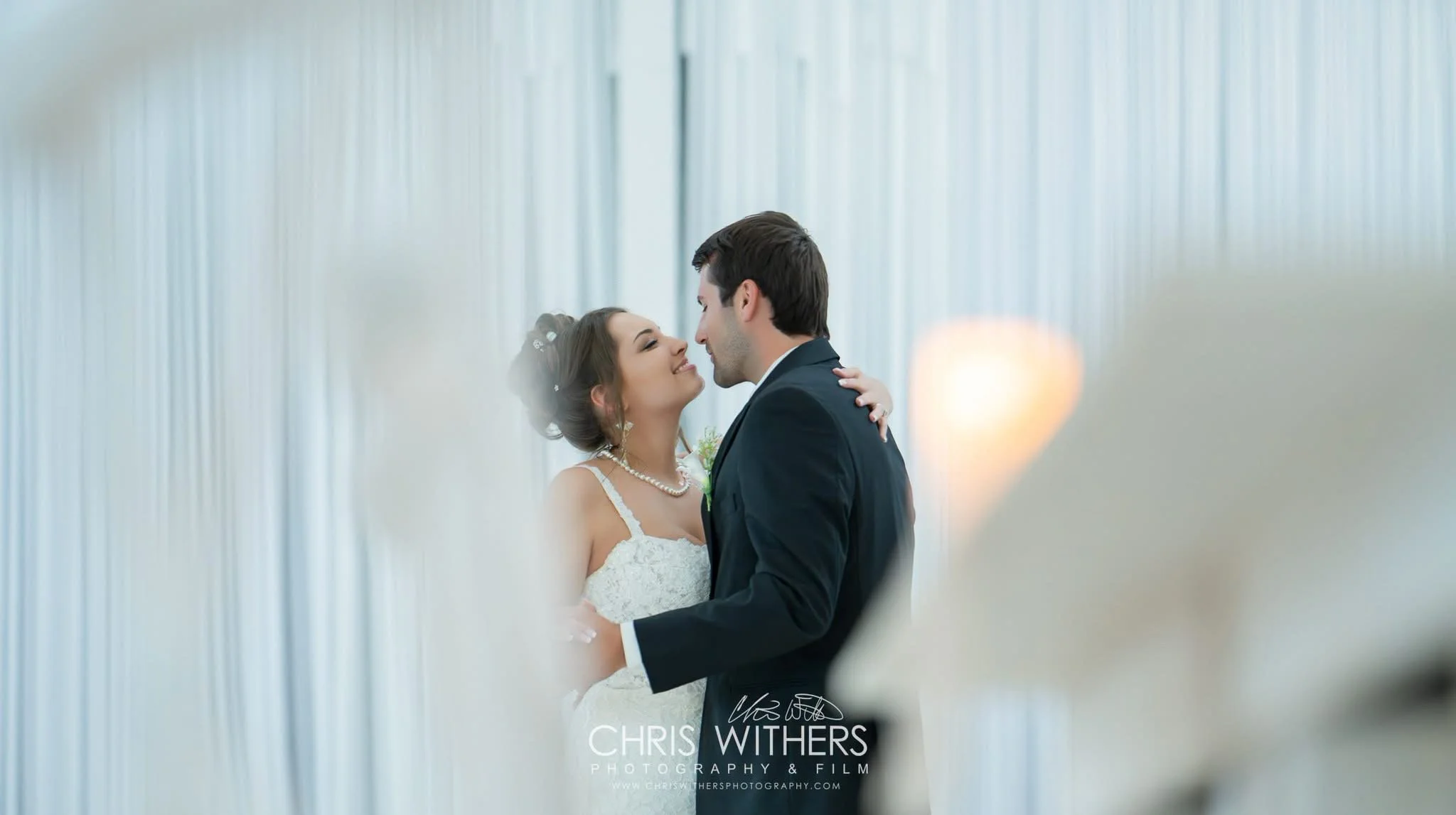 A bride and groom sharing a close moment during their wedding in front of a striped white curtain, smiling at each other.