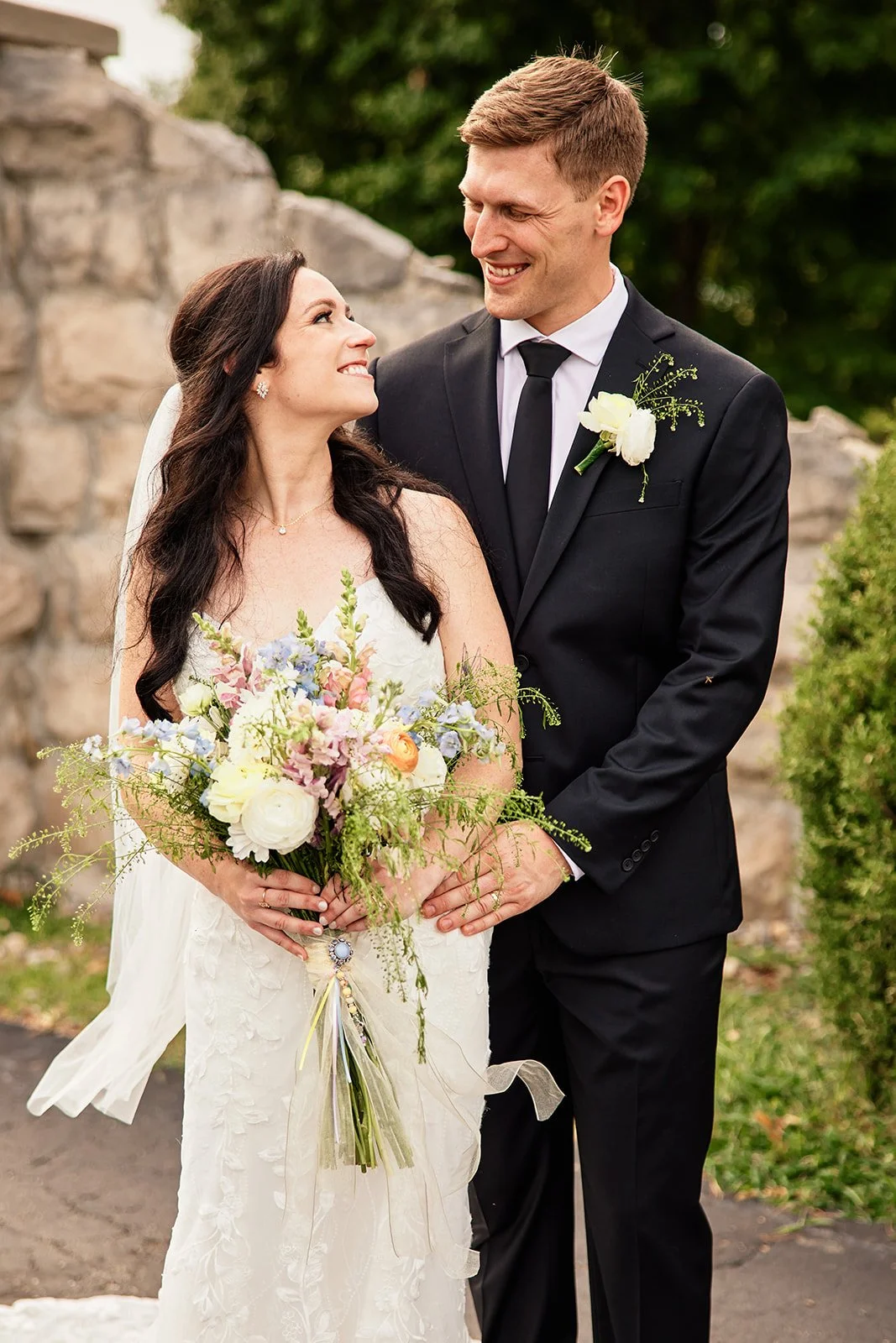 A bride and groom smiling at each other outdoors, with the bride holding a bouquet of flowers and wearing a white wedding dress, and the groom in a black suit with a white boutonniere, standing in front of a stone wall with greenery.