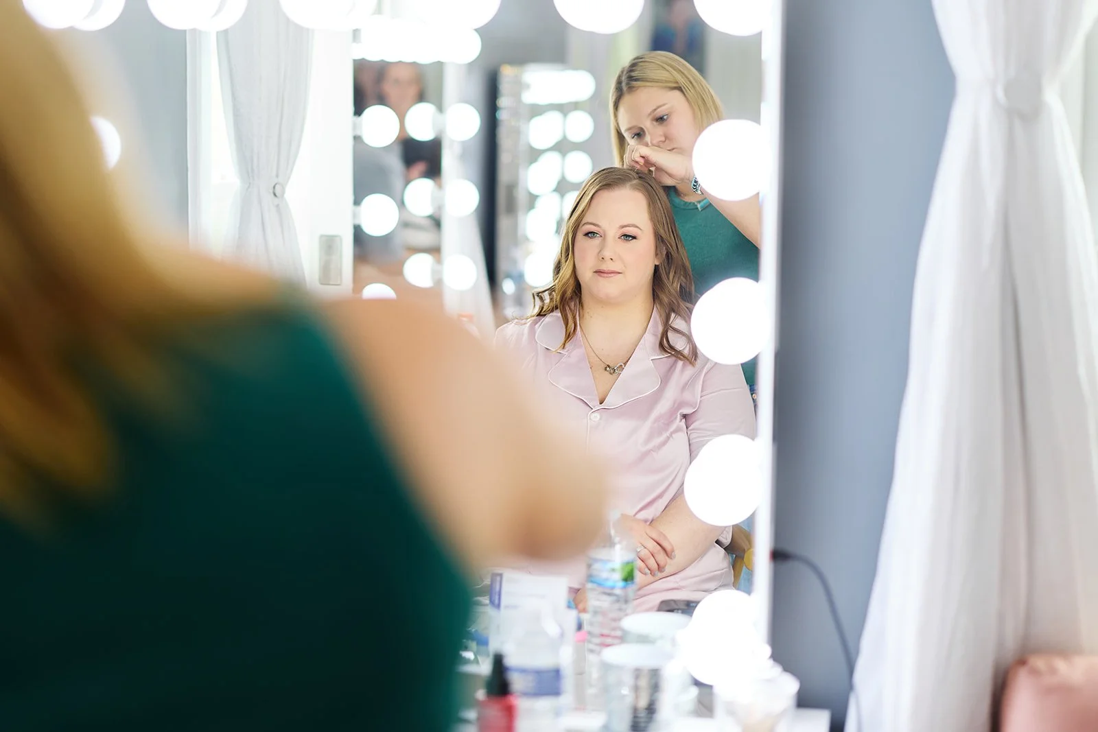 A woman is sitting in front of a mirror with makeup and beauty products on the table. A makeup artist is standing behind her, working on her hair. The mirror has round, illuminated light bulbs around its frame.