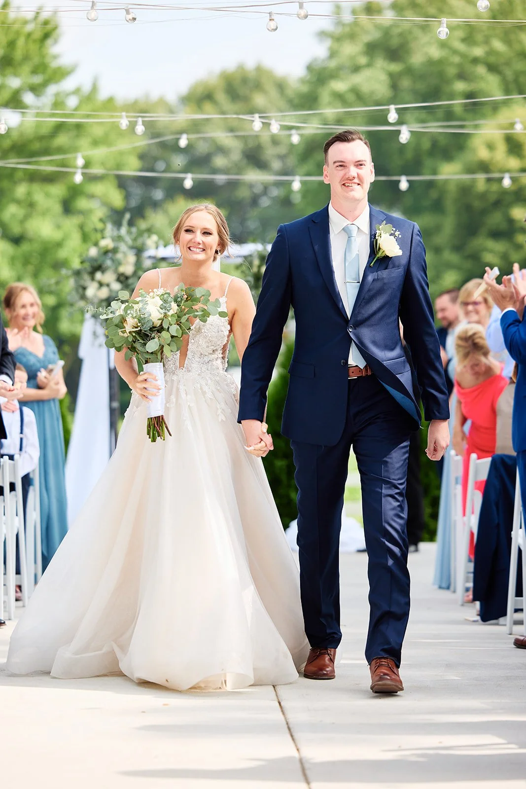 A bride and groom smiling and holding hands as they walk down the aisle at an outdoor wedding ceremony, surrounded by guests.