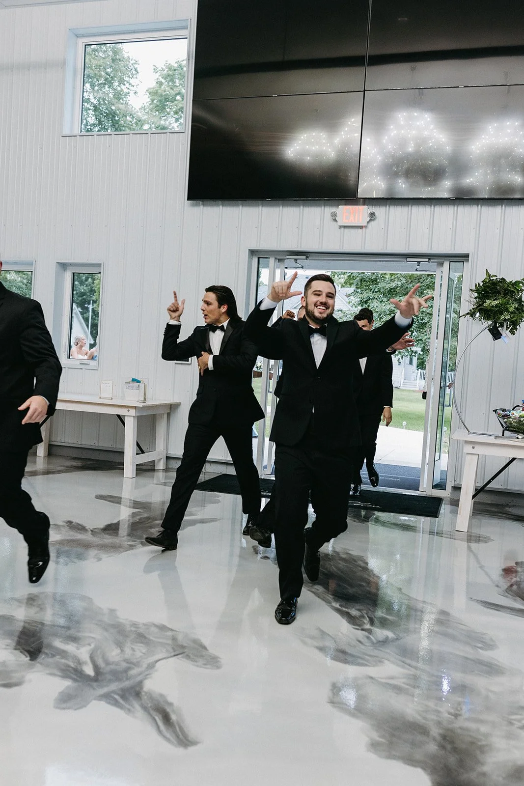 A group of men dressed in tuxedos dancing and smiling in a modern event space with white walls, large windows, and a polished concrete floor.