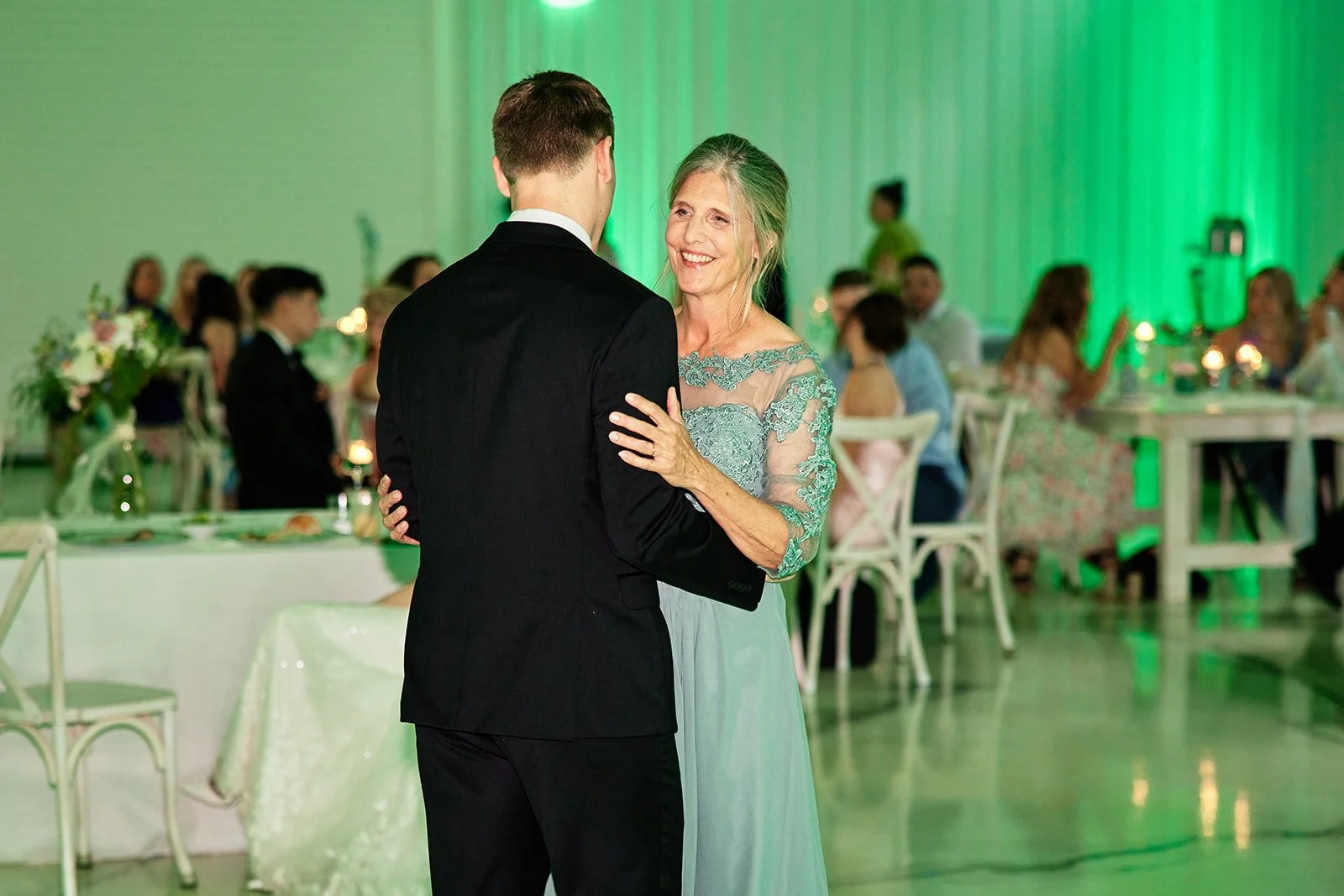 A woman and a man dancing at a wedding reception, with guests seated at tables in the background illuminated by green lighting.