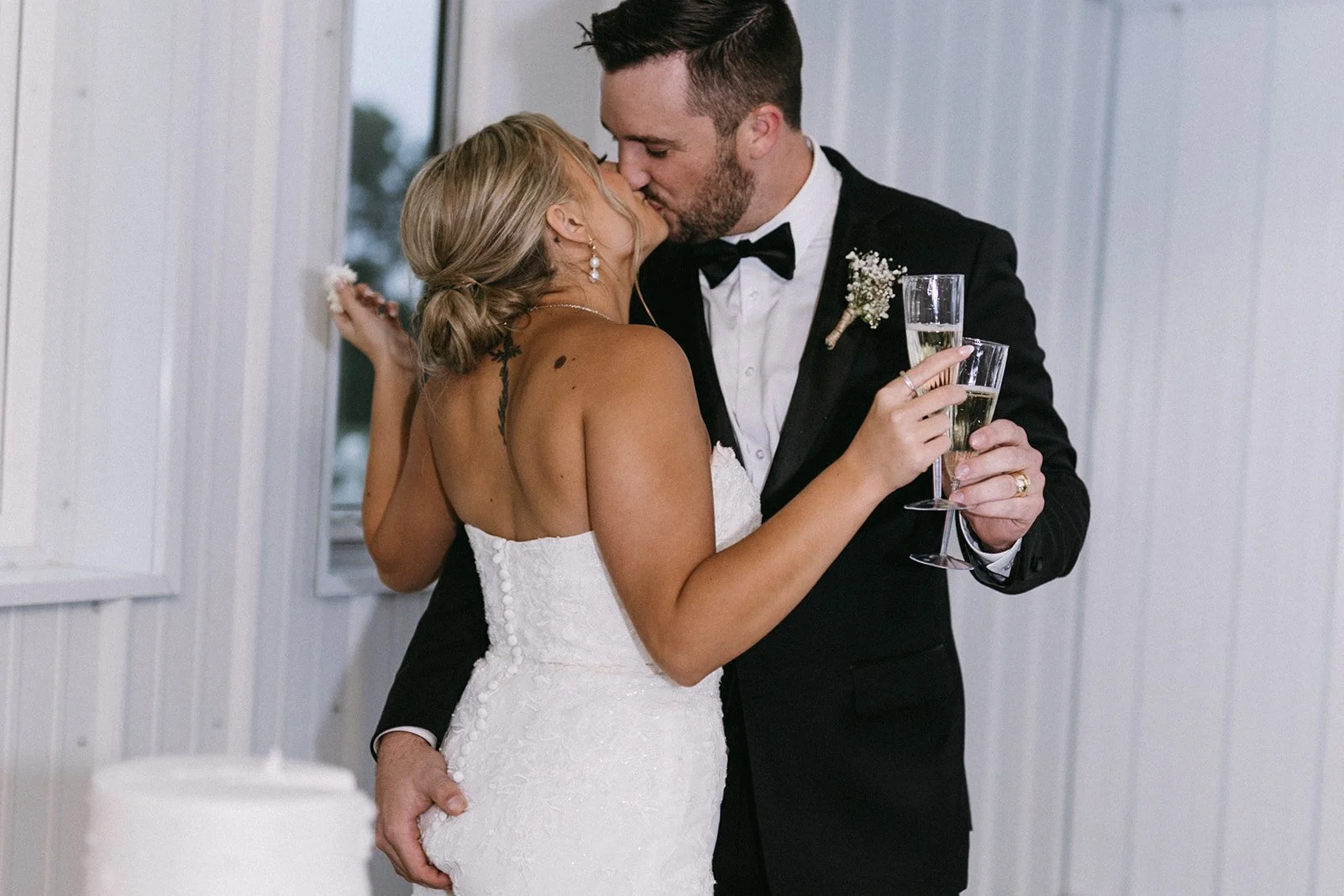 A newlywed couple sharing a kiss and dancing, with the bride holding two glasses of champagne, in a room with white walls and windows.