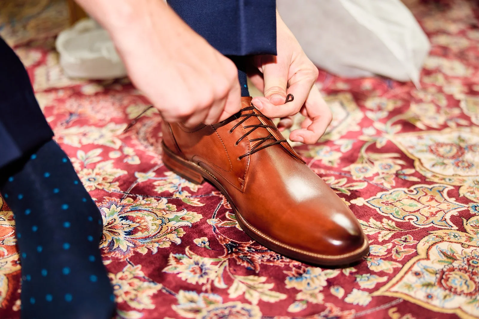 Person tying the laces of a brown leather dress shoe on a patterned red area rug.