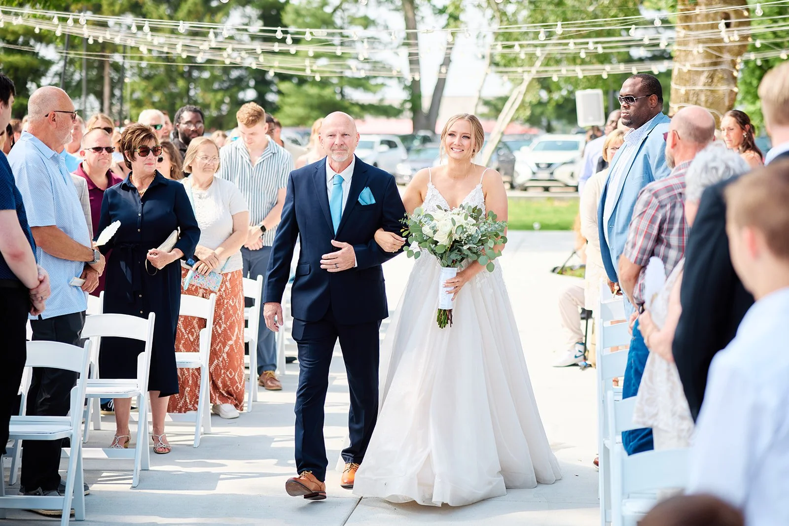 Bride walking down the aisle with her father at an outdoor wedding ceremony, surrounded by seated guests and string lights overhead.