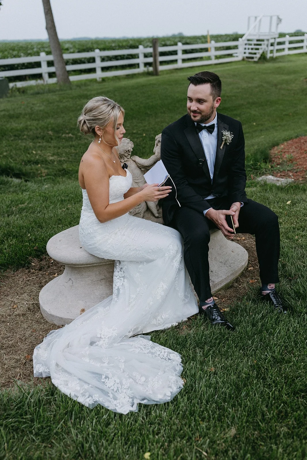 A bride and groom sitting on a stone bench outdoors, with the bride reading from a notebook and the groom holding a phone, in wedding attire on a grassy area near a white fence.