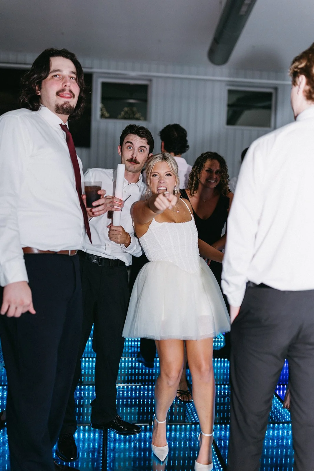 Group of people at a party, with a woman in a white dress pointing and angry, while others look surprised or amused.