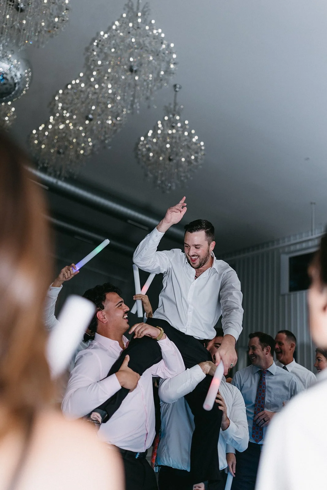 Group of people celebrating at a party, with one man being lifted on another's shoulders, holding glow sticks, under chandeliers.