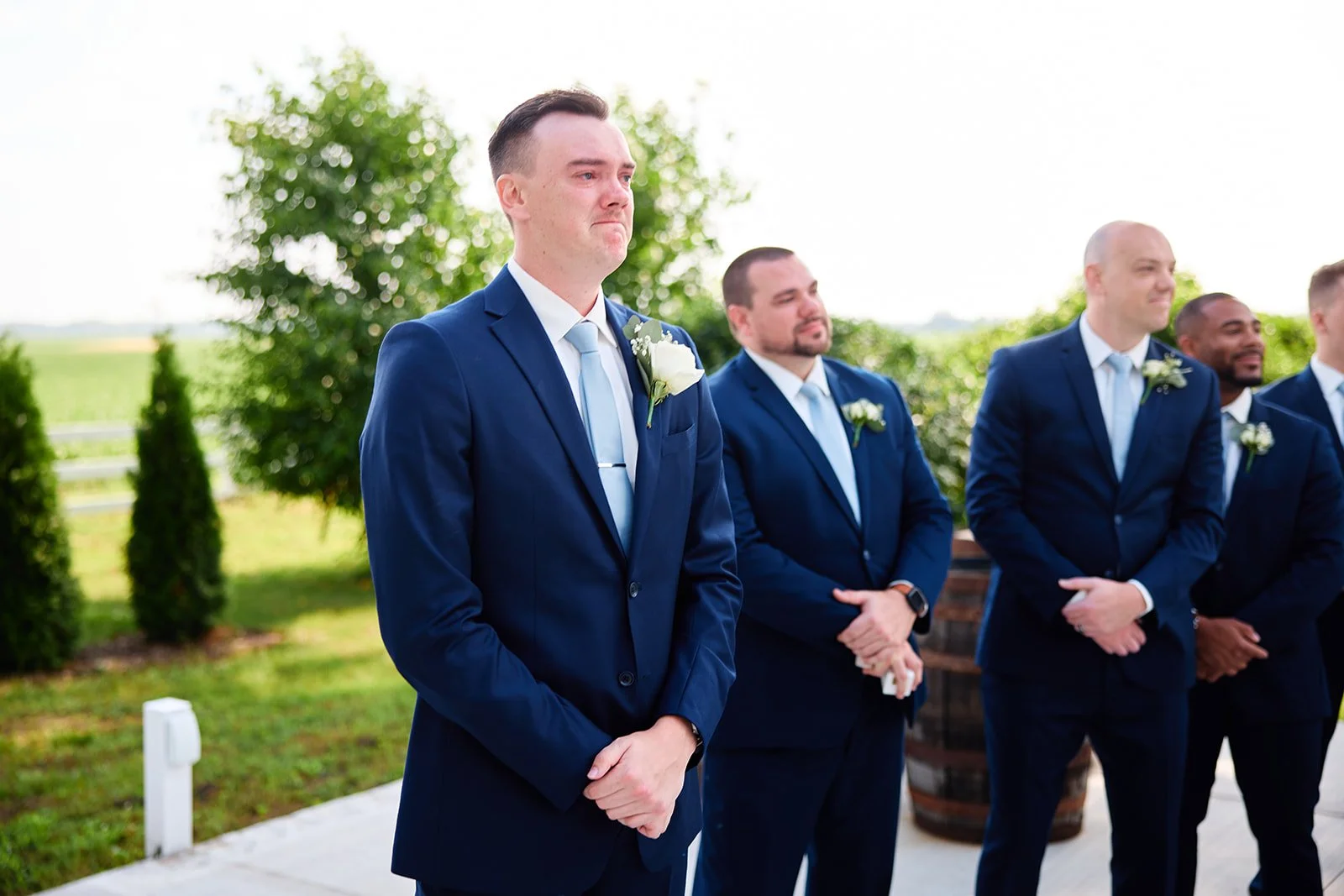 A groom and groomsmen in blue suits standing outdoors during a wedding ceremony, with trees and open sky in the background.