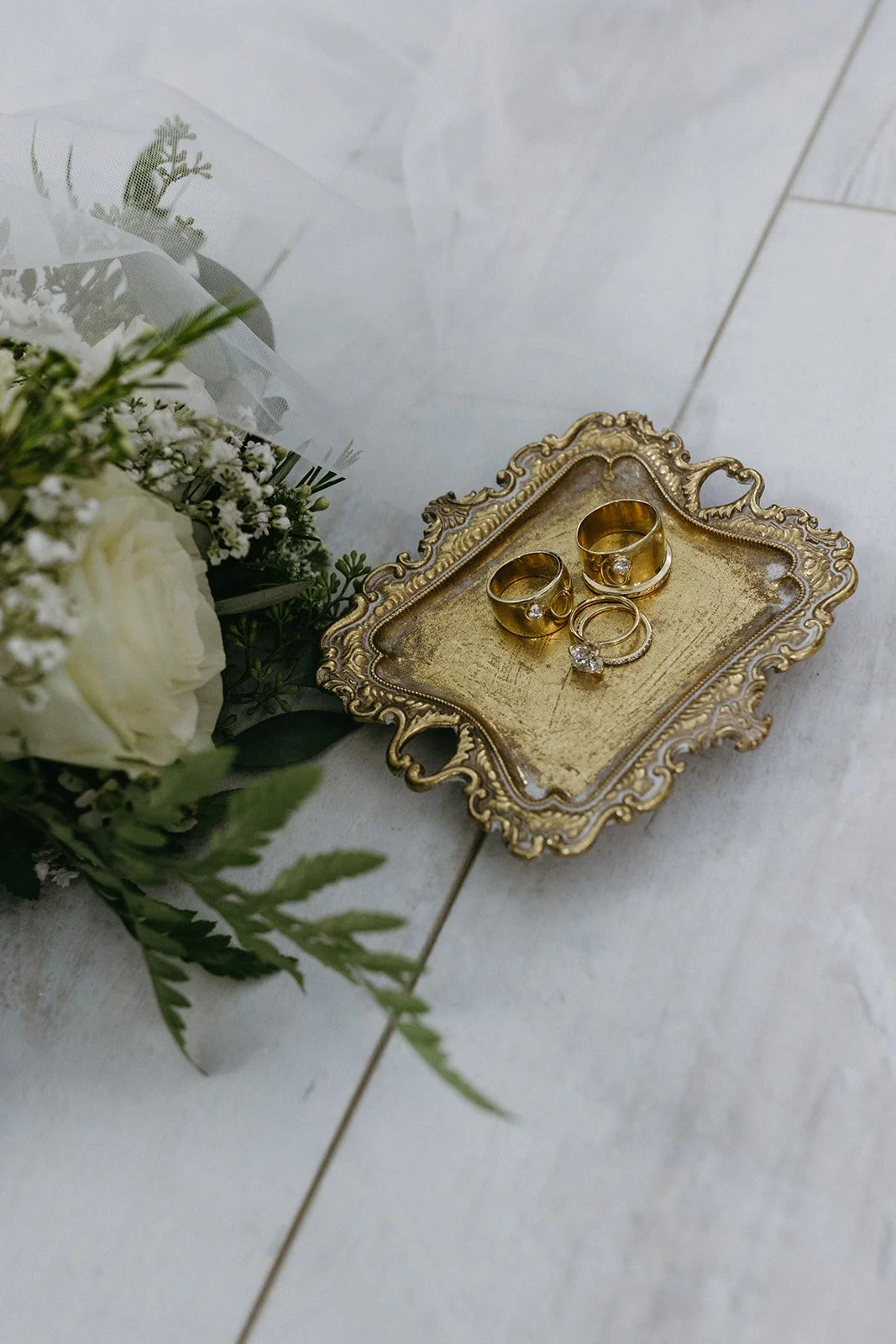 Wedding rings, including two gold bands with diamonds and a diamond engagement ring, on an ornate gold tray beside a bouquet of white roses and greenery.