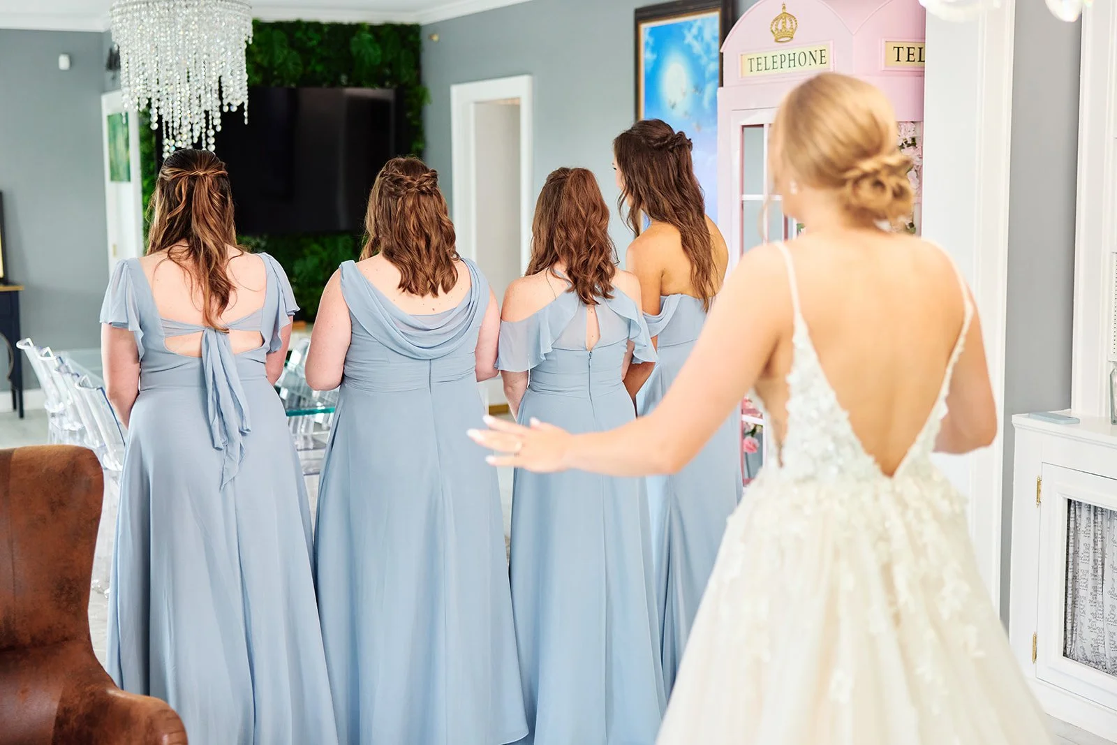 A bride in a white wedding dress with a low back and lace details reaching out to her bridesmaids, who are dressed in matching light blue dresses, in a bright decorated room with a pink telephone booth prop in the background.
