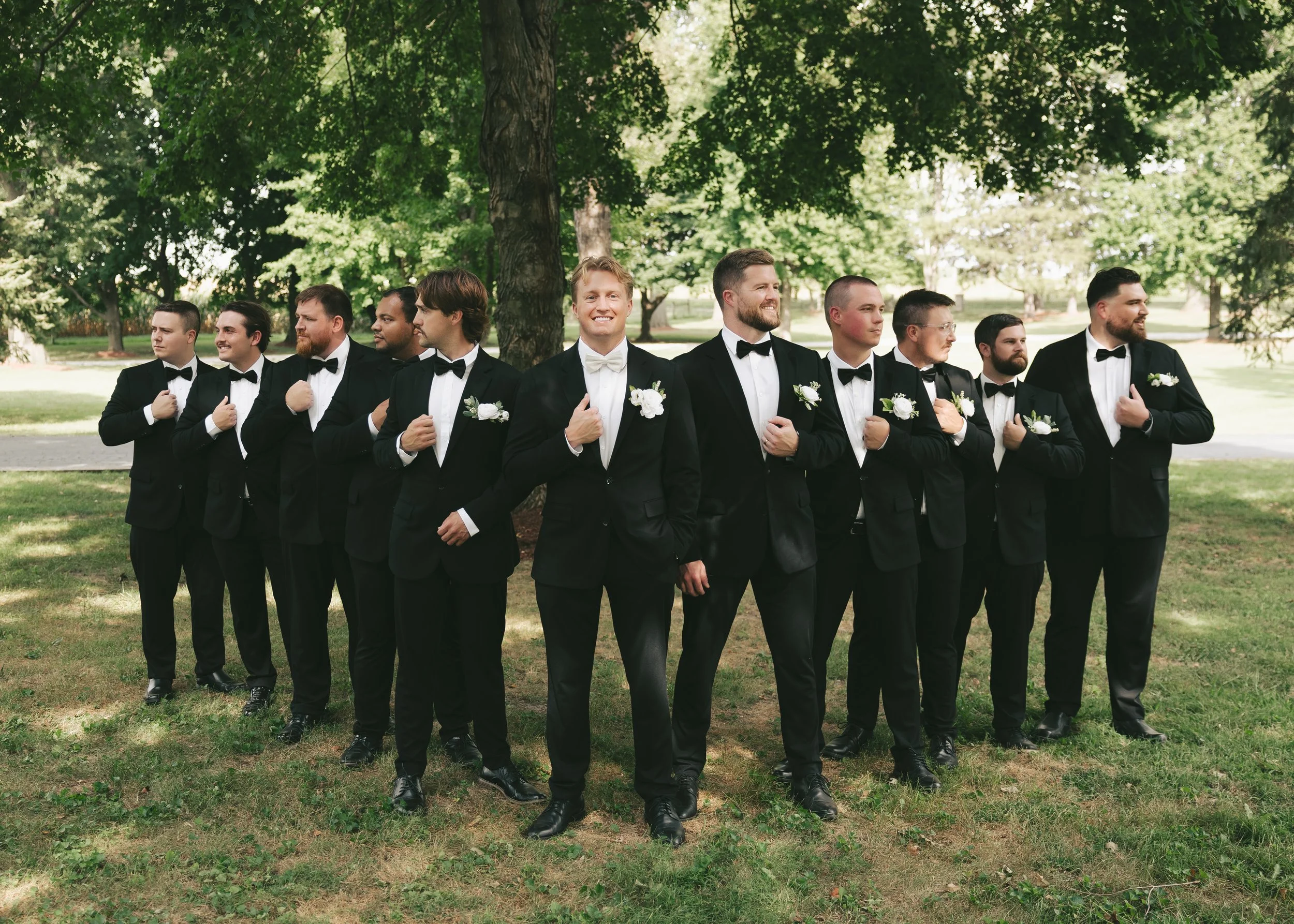 A group of men dressed in tuxedos with black bow ties and white shirts, holding each other's shoulders or arms, standing in a line outdoors at a wedding venue in Springfield, ILwith green grass and lush trees. Pole Barn Chic.