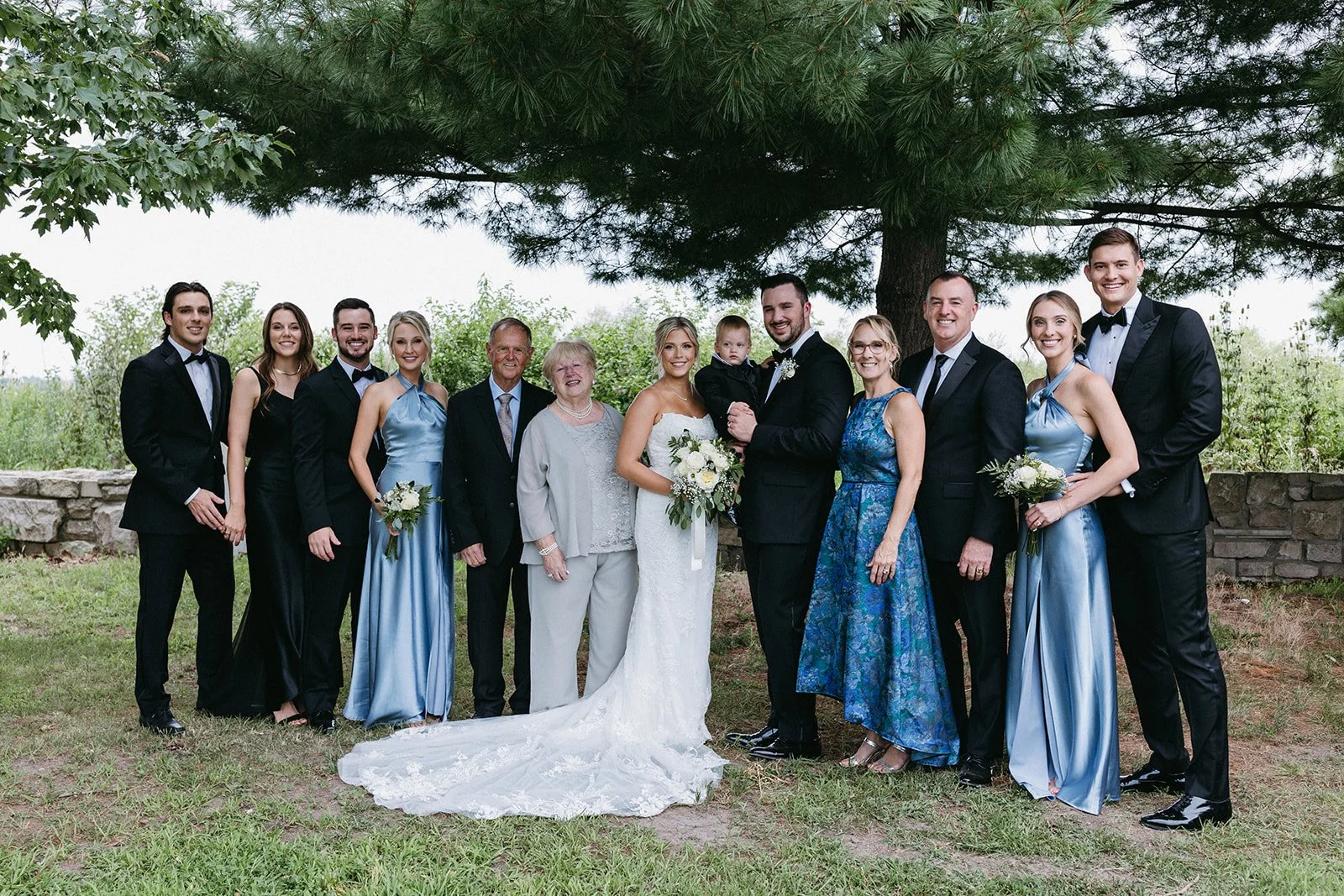 A large wedding party of 13 people, including bride and groom, standing outdoors under a large tree with greenery and stone wall in the background. The bride is in a white wedding dress holding a bouquet of white roses, and the groom is in a black tu