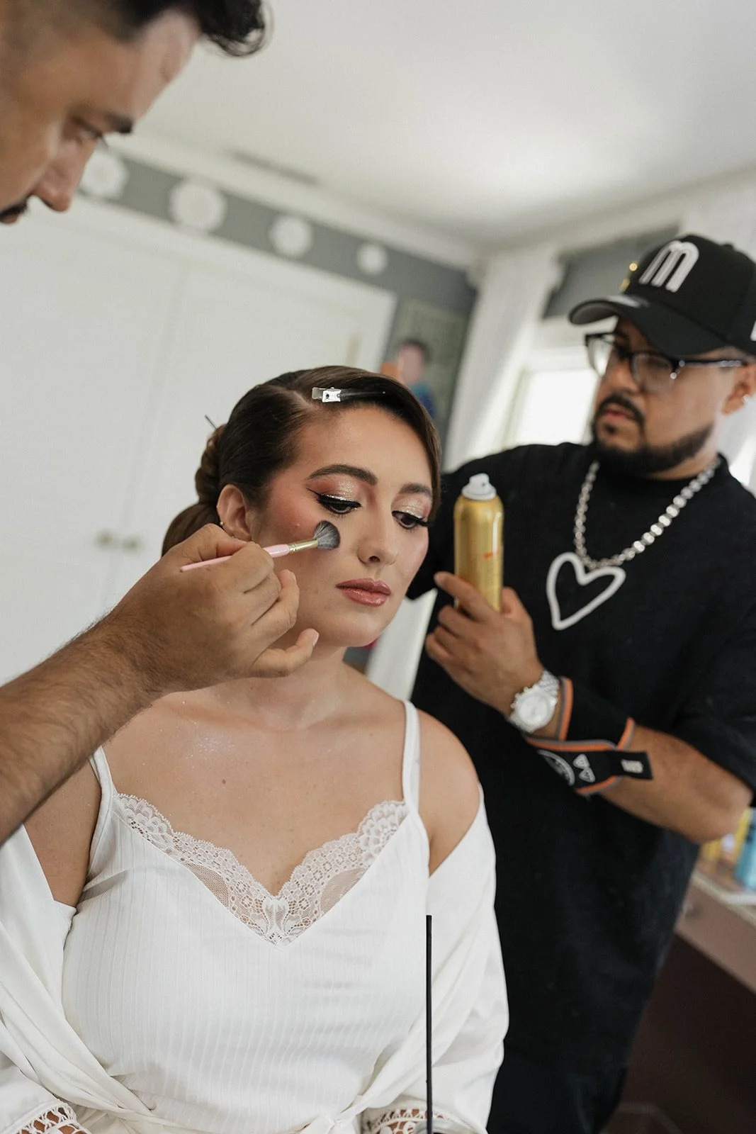 A woman with a white top and lace details getting her makeup done by a makeup artist, with another man holding a can of makeup spray in the background.