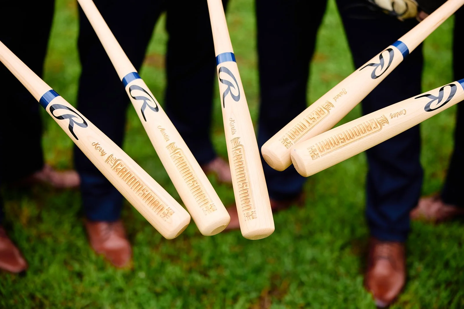 A group of baseball bats being held in a circle, with the tips pointing toward the center, against a background of grass and people wearing dark pants and brown shoes.