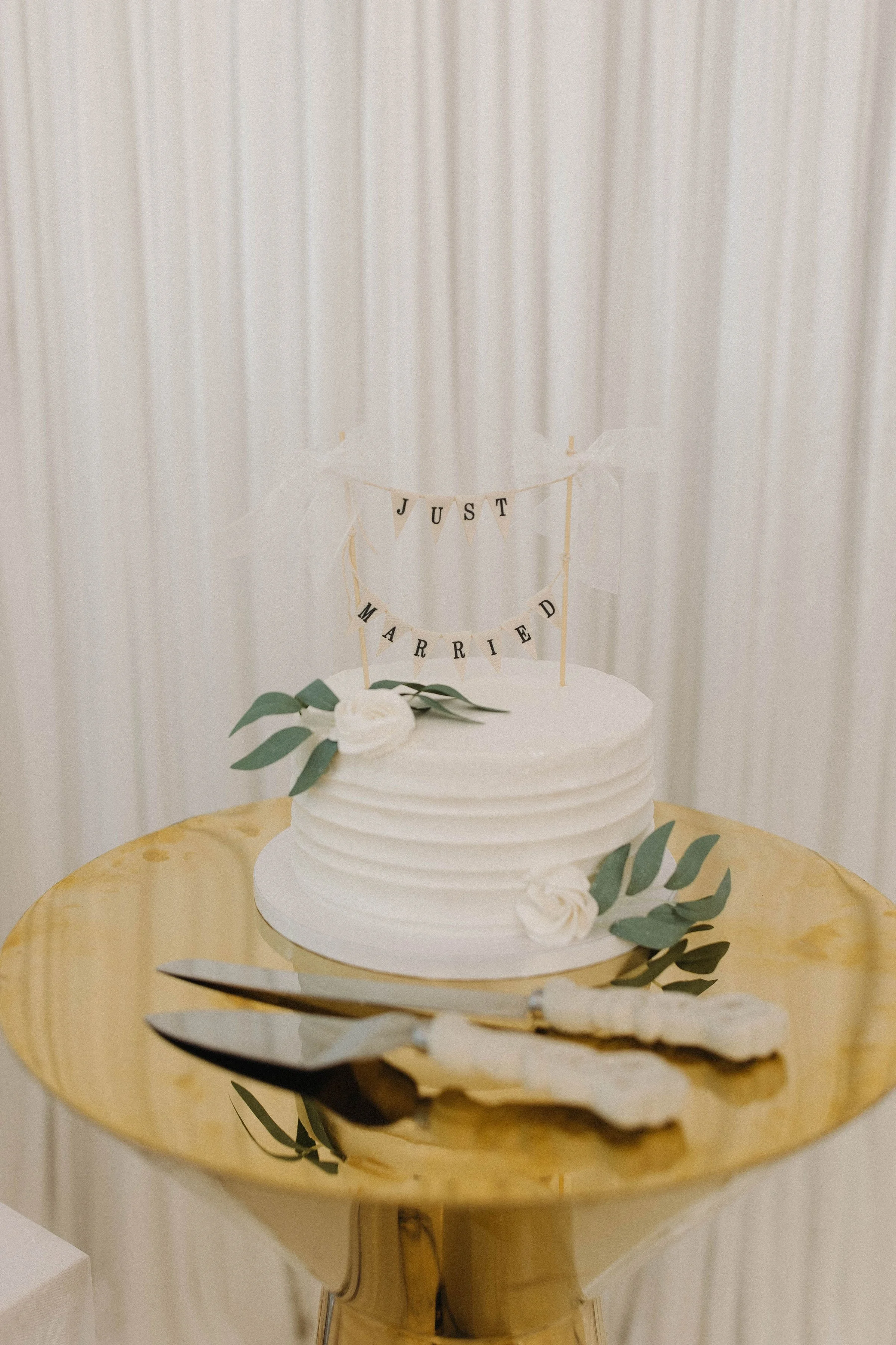 White wedding cake with green leaves and white roses on a gold cake stand, topped with a 'Just Married' banner. Silver cake servers rest on the stand, set against a neutral background.