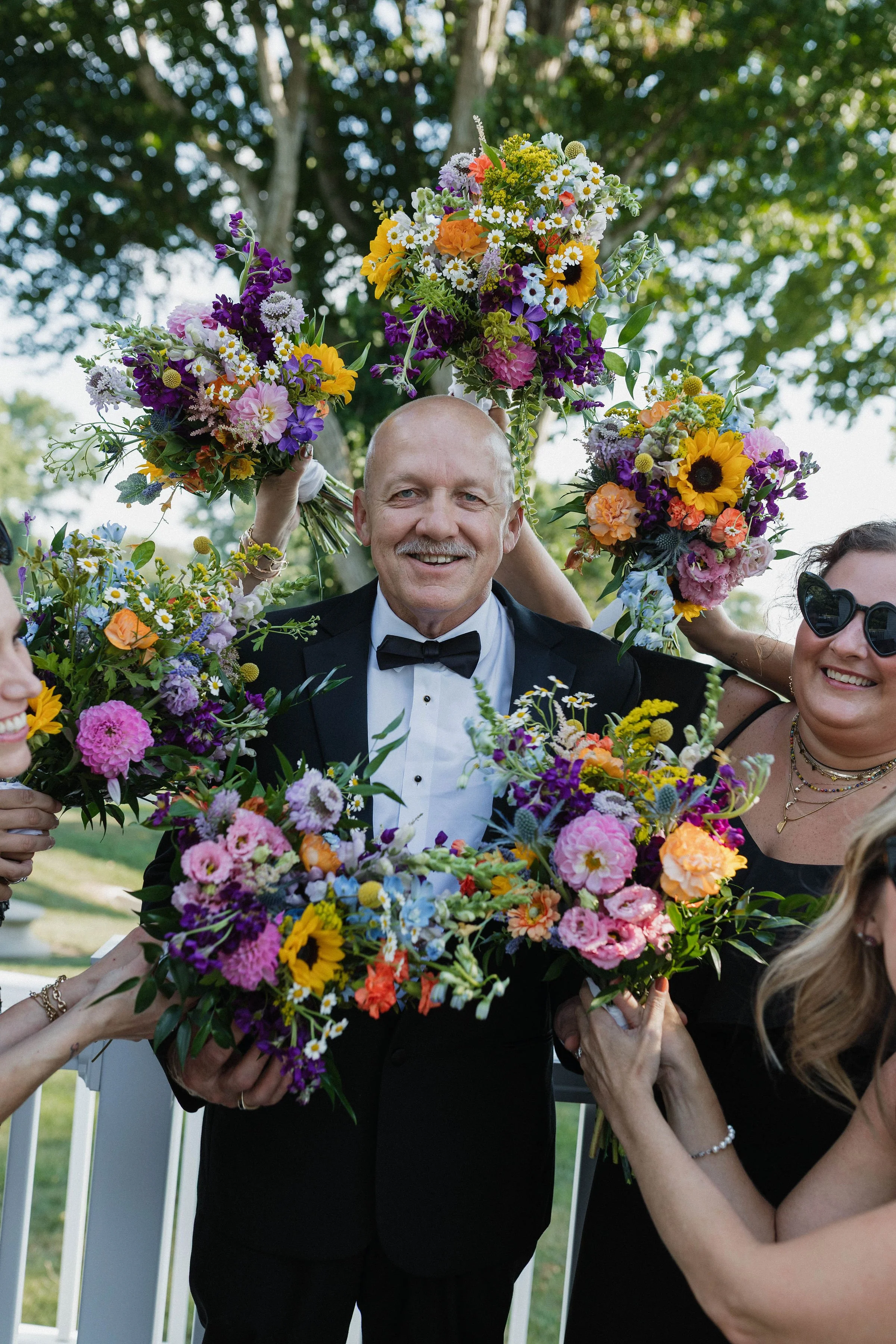 A man in a tuxedo holding multiple colorful flower bouquets during an outdoor celebration surrounded by smiling women.