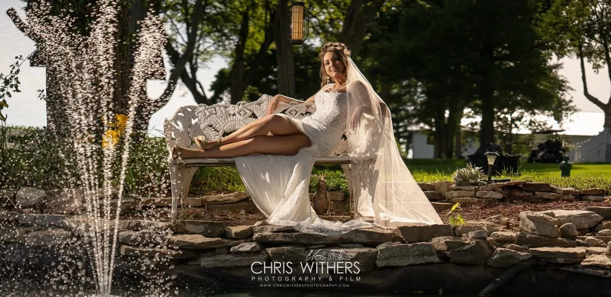 A woman in a wedding dress and veil relaxing on a decorative bench beside a pond with a fountain in a park setting.