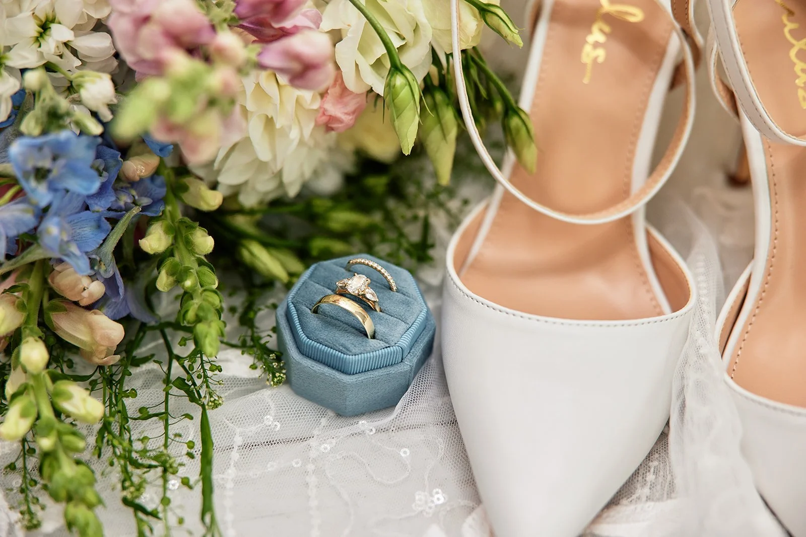 Wedding rings on a blue ring box with flowers, white high-heeled shoes, and light pink ballet flats.