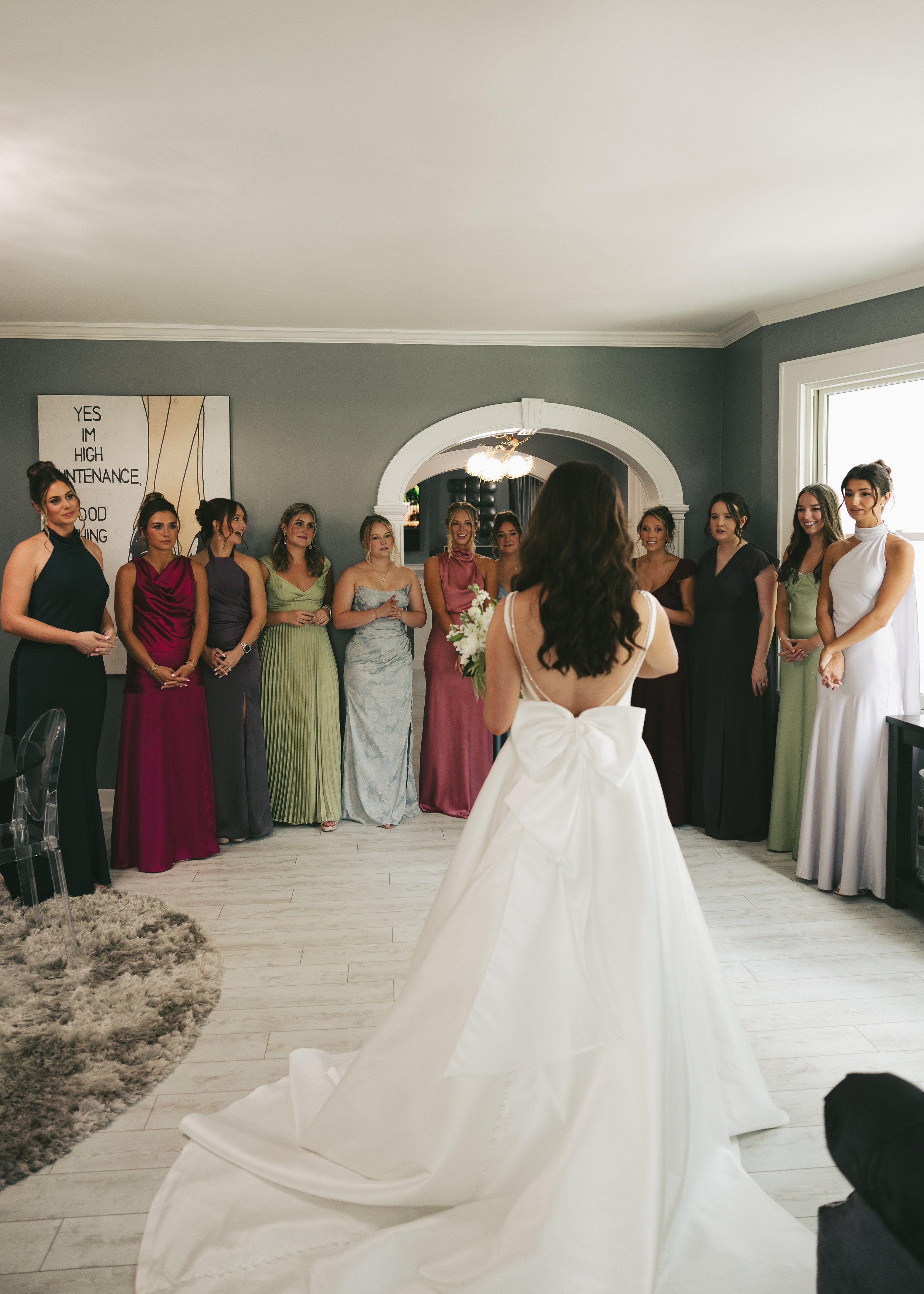 A bride in a white wedding dress with a large bow at the back stands facing a group of women in colorful dresses, who are watching and smiling. The women are in a living room with gray walls, a large window, and a framed text art on the wall.