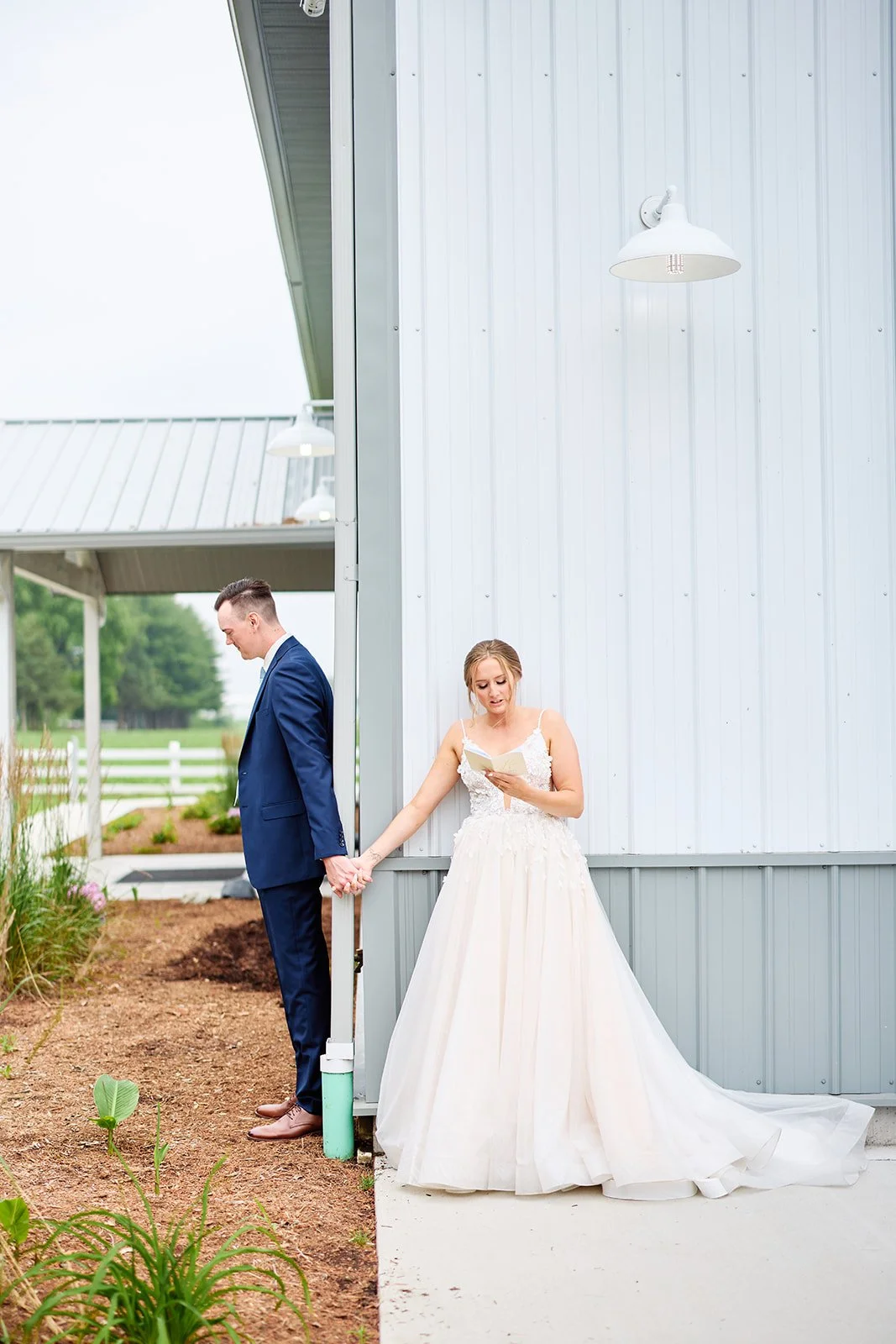 A bride and groom holding hands outside a white building, with the bride reading a card or letter. The groom is dressed in a navy suit, and the bride in a white wedding gown.