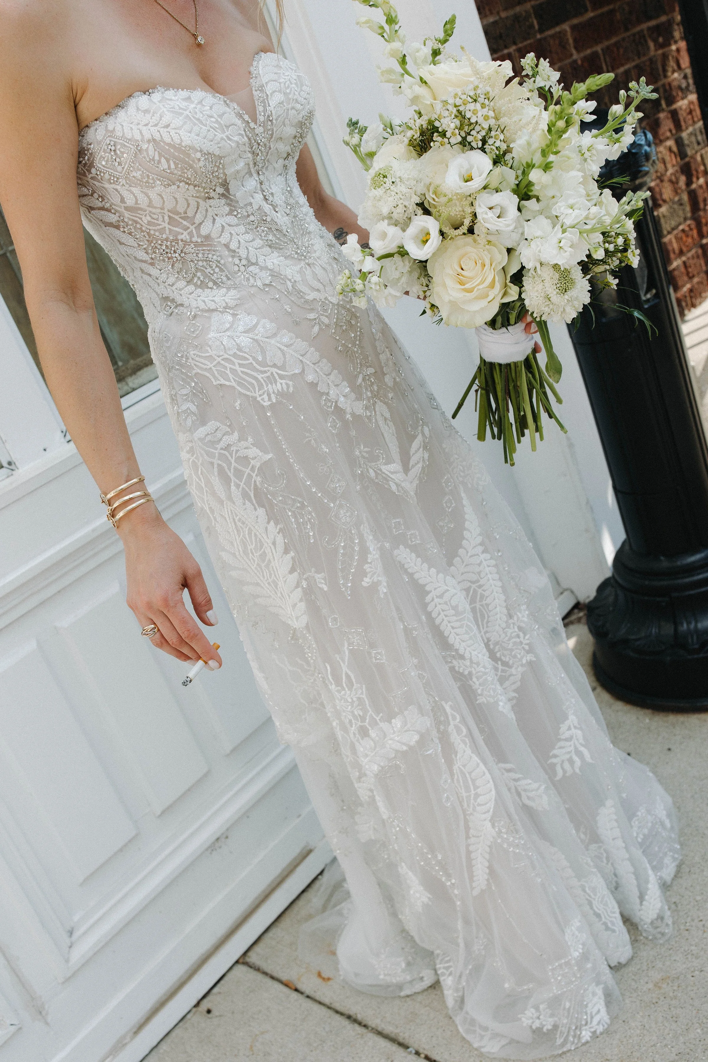 A bride in a white dress with intricate lace and beadwork holding a bouquet of white flowers, with a cigarette in her left hand, standing outside near a white door and brick wall.