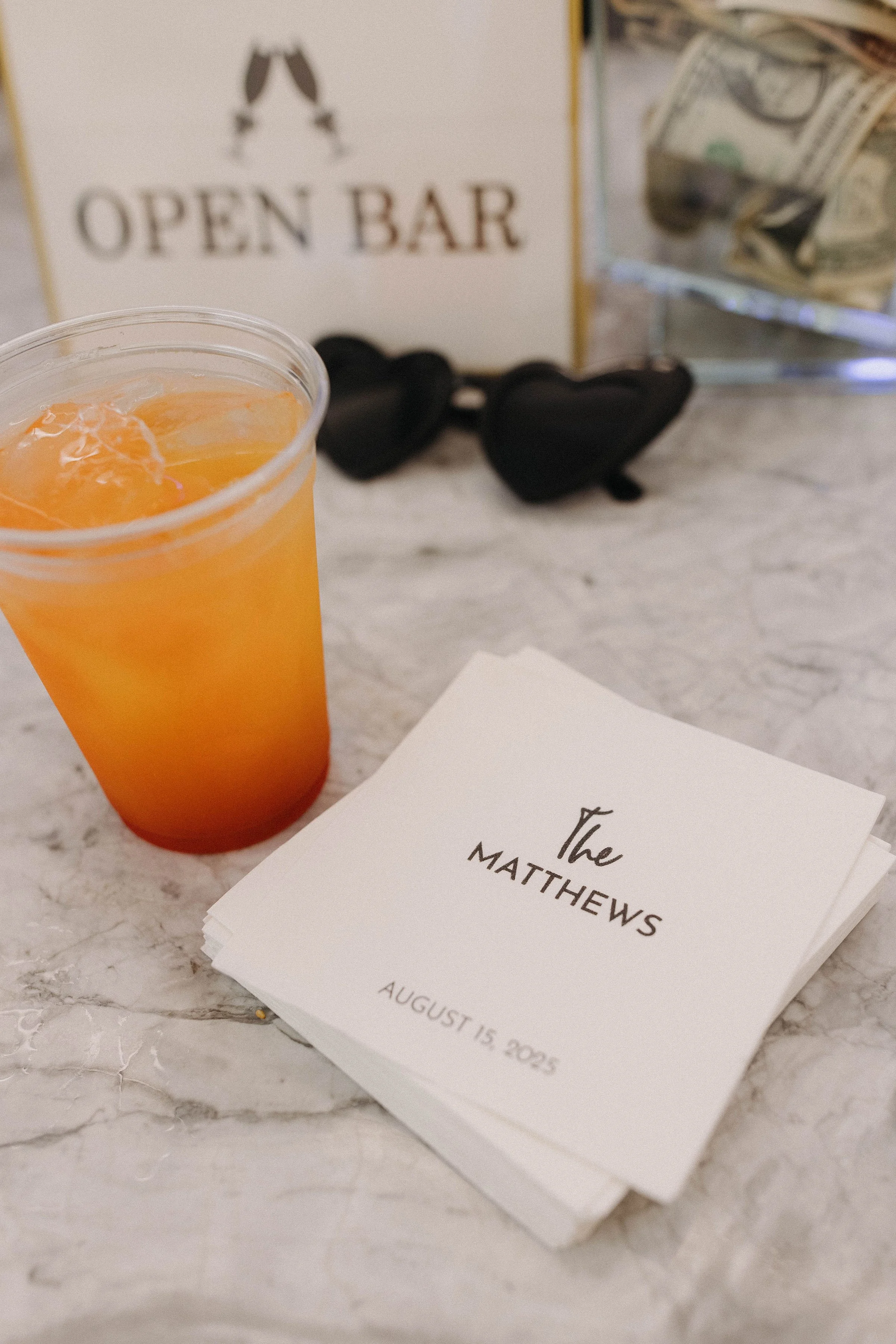Wedding reception table with a drink, a stack of place cards, black heart-shaped sunglasses, and a sign that says 'Open Bar' with a pair of clinking glasses illustration. 