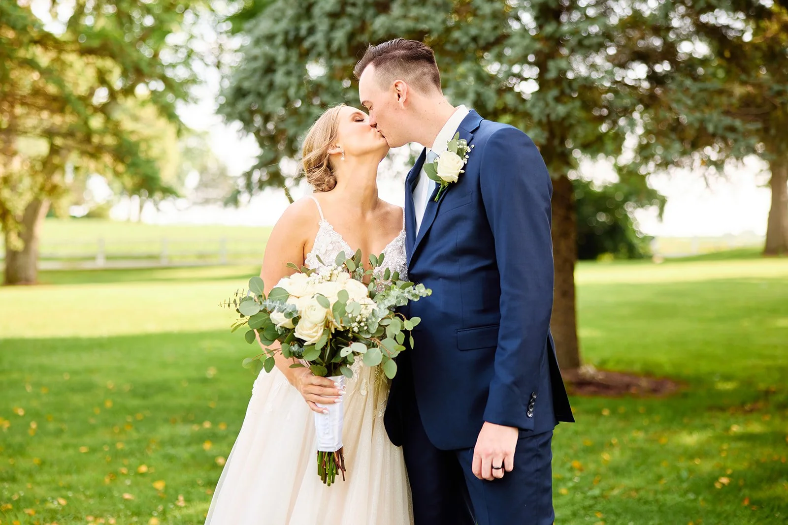 A bride and groom sharing a kiss outdoors on their wedding day, surrounded by trees and grass, with the bride holding a large bouquet of white roses and greenery.