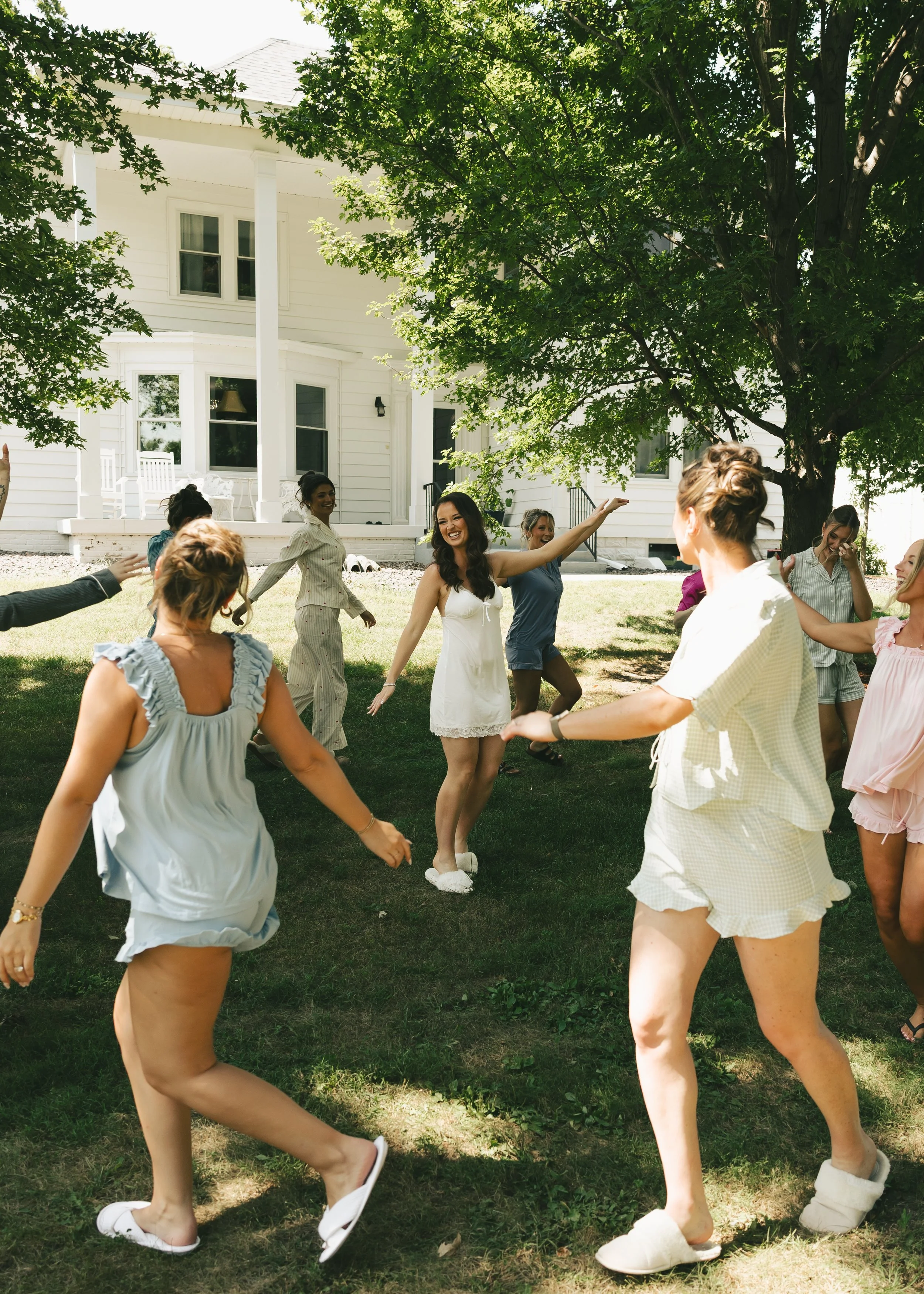 A group of women dancing and celebrating outdoors on a grassy lawn next to a white house with large trees providing shade. The women are wearing summer dresses and appear happy and joyful.