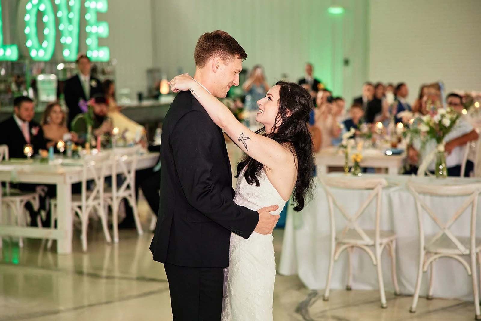 Couple dancing at wedding reception, with guests seated at decorated tables in background.