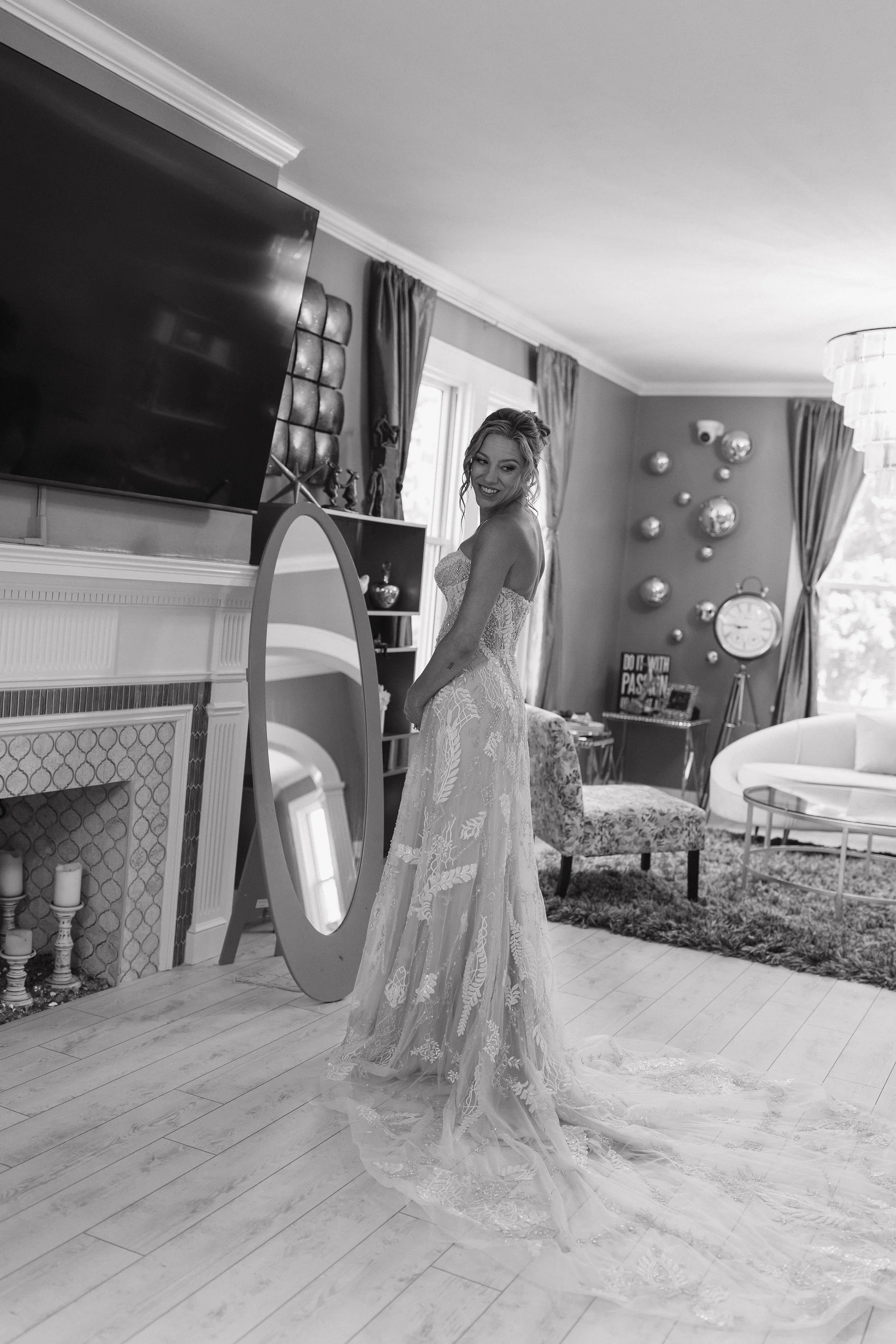 A woman in a wedding dress standing in a living room, smiling, with a large mirror, television, and modern decor in the background.