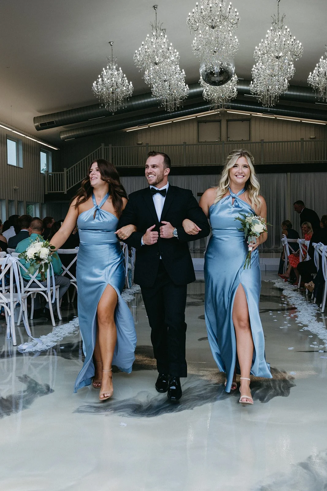 A groom walking down the aisle with two bridesmaids at a wedding reception, with chandeliers hanging from the ceiling and guests seated on either side.