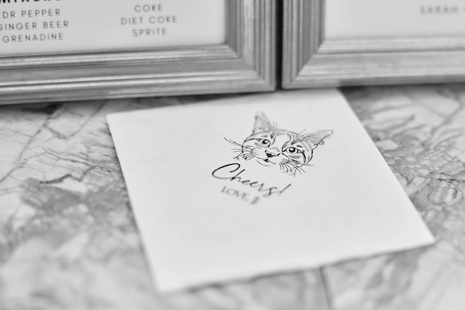 A greeting card with an illustration of a cat's face and the words 'Cheers! Love, J' on a marble countertop, with a paper towel dispenser in the background.