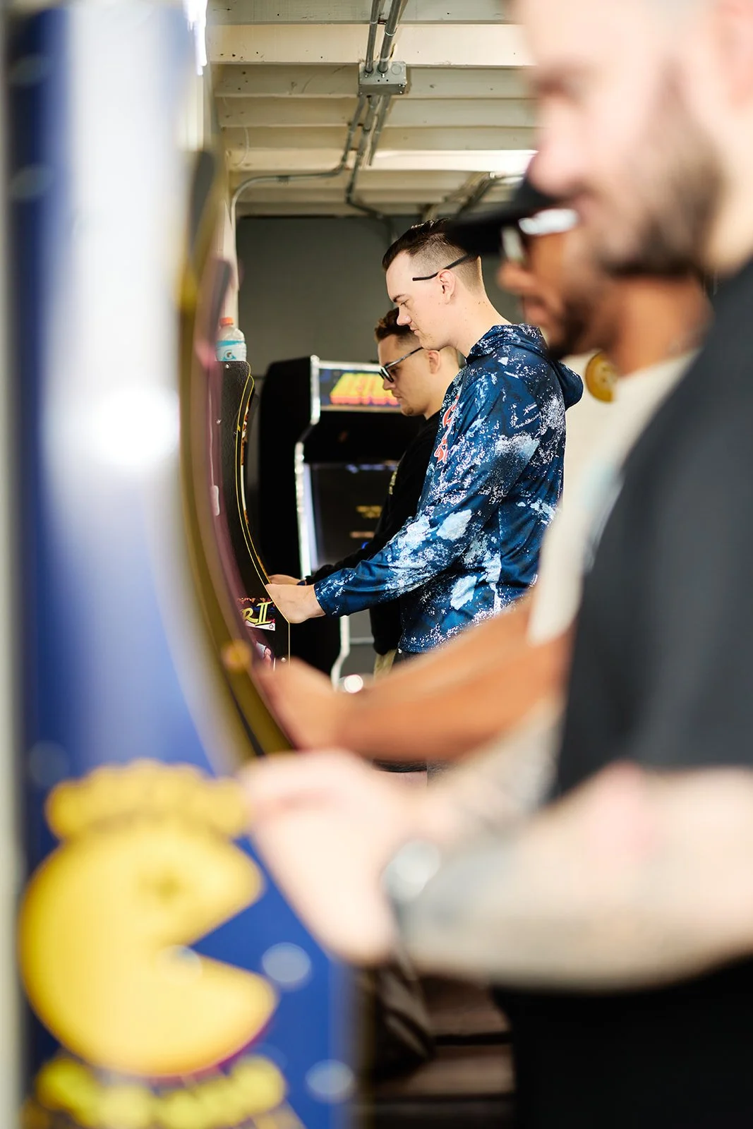 Three young men playing arcade video games in a row at an arcade, with focus on the middle player wearing a blue galaxy patterned hoodie.