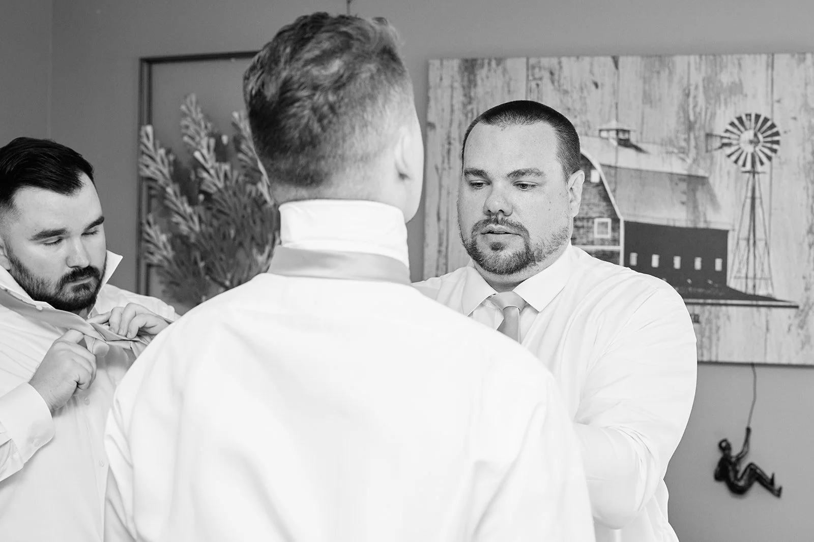 Three men in white dress shirts getting ready, one fixing his tie, in a room with farm-themed wall art and a hanging figure on the wall.