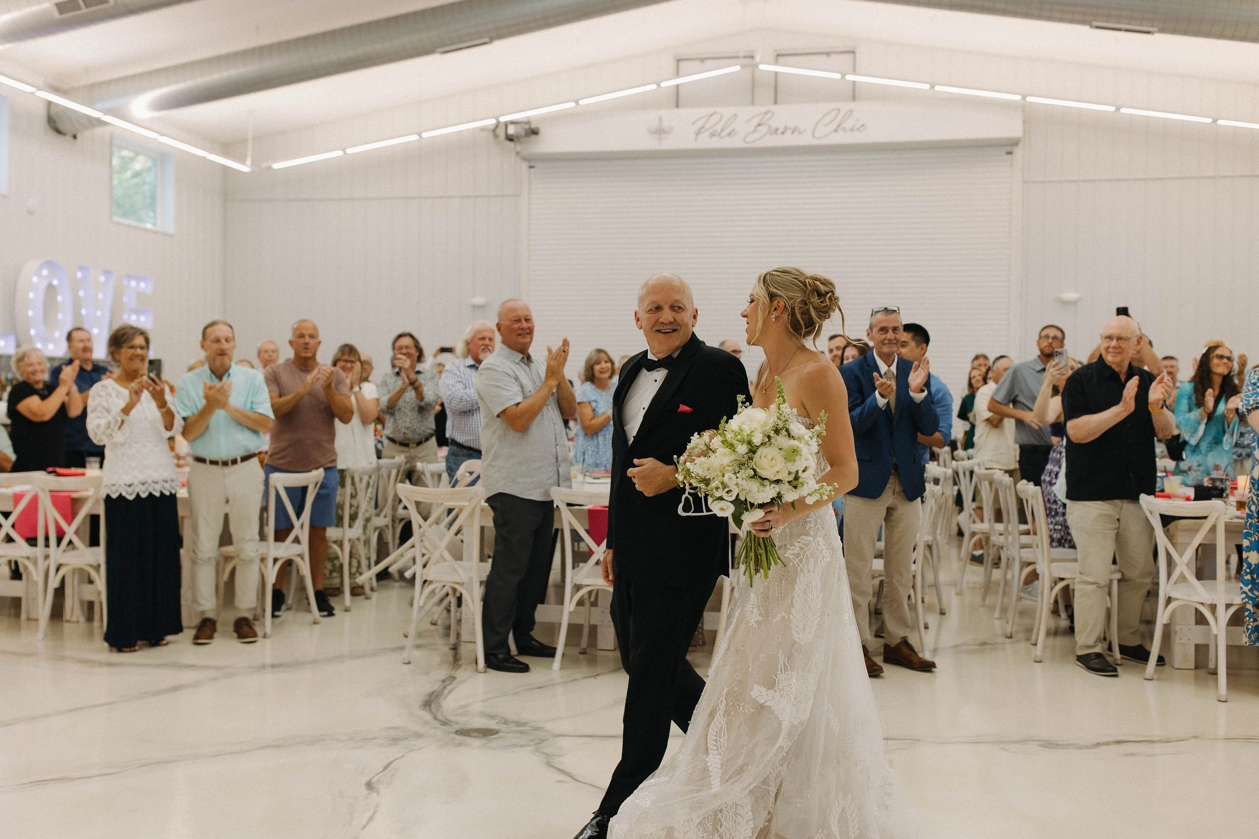 Bride walking down the aisle with her father, holding a bouquet, at a wedding reception in a bright, white hall with guests clapping and smiling.