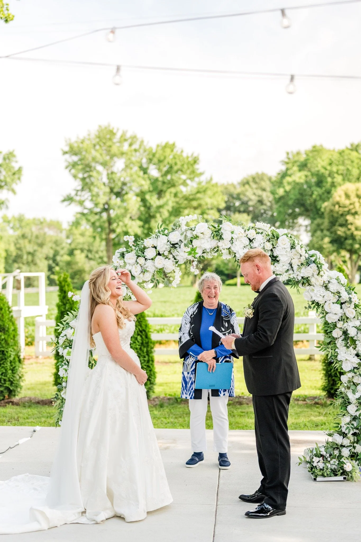 A bride and groom exchanging vows under a floral arch at an outdoor wedding ceremony, officiant standing behind them, with green trees and string lights overhead.