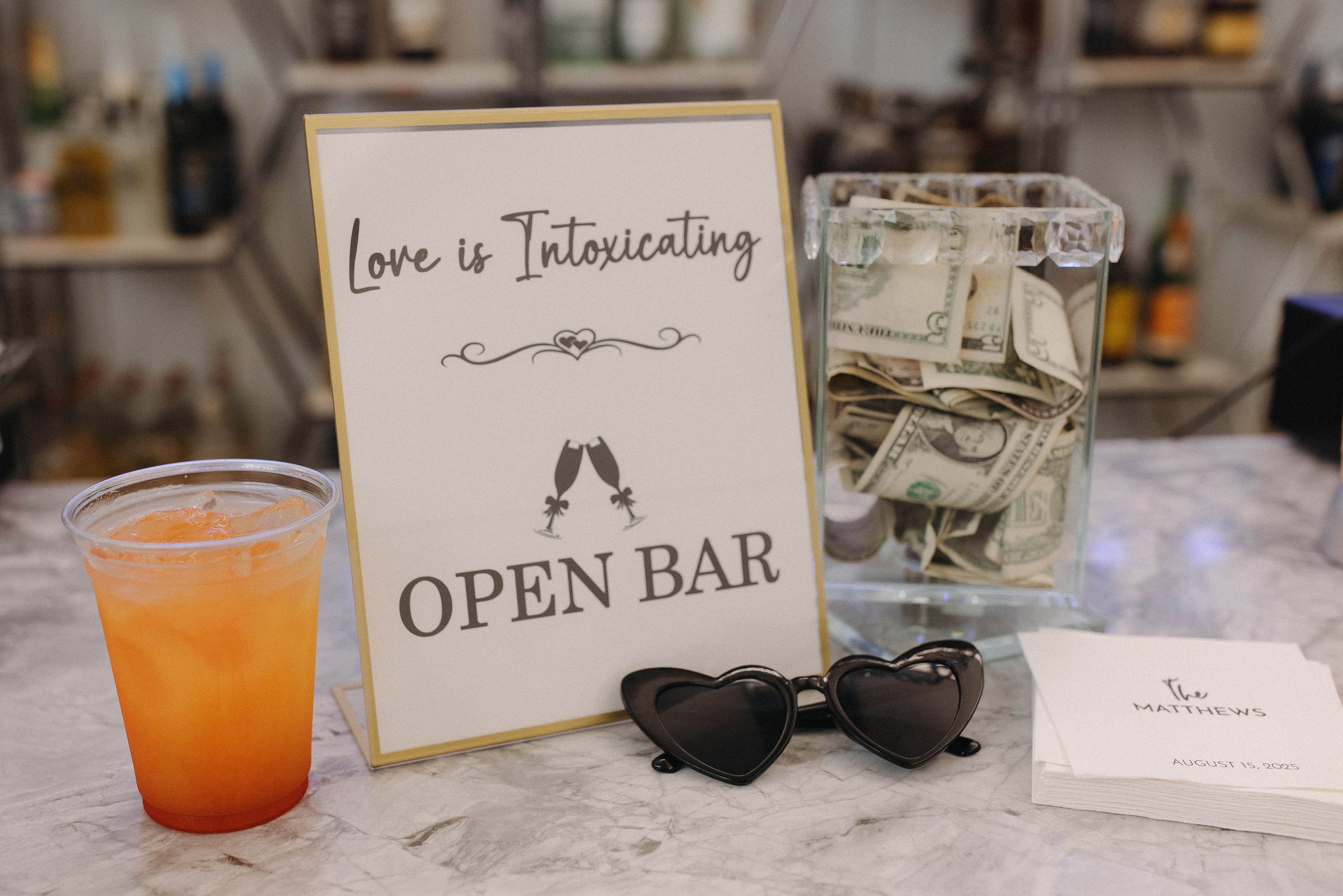 Table display with a drink, a sign reading 'Love is intoxicating, open bar,' a cash jar filled with dollar bills, sunglasses, and a stack of napkins with printed text.