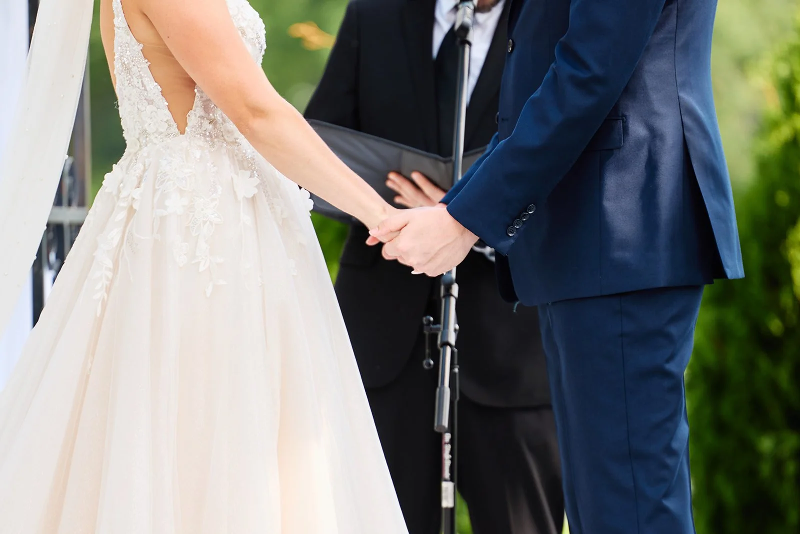 A bride and groom holding hands during their outdoor wedding ceremony, with an officiant standing in between them.